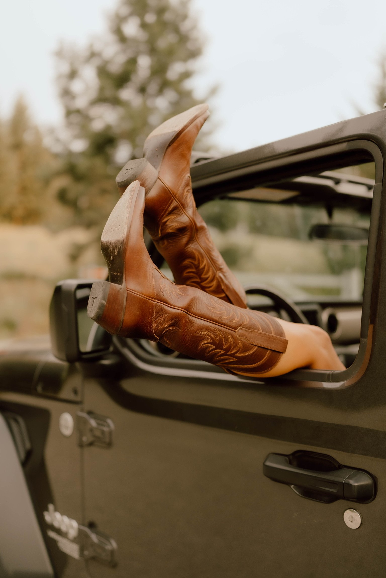 A person wearing brown leather shoes is seen stretching their foot outside a vehicle with the door open. The background shows trees and a cloudy sky.