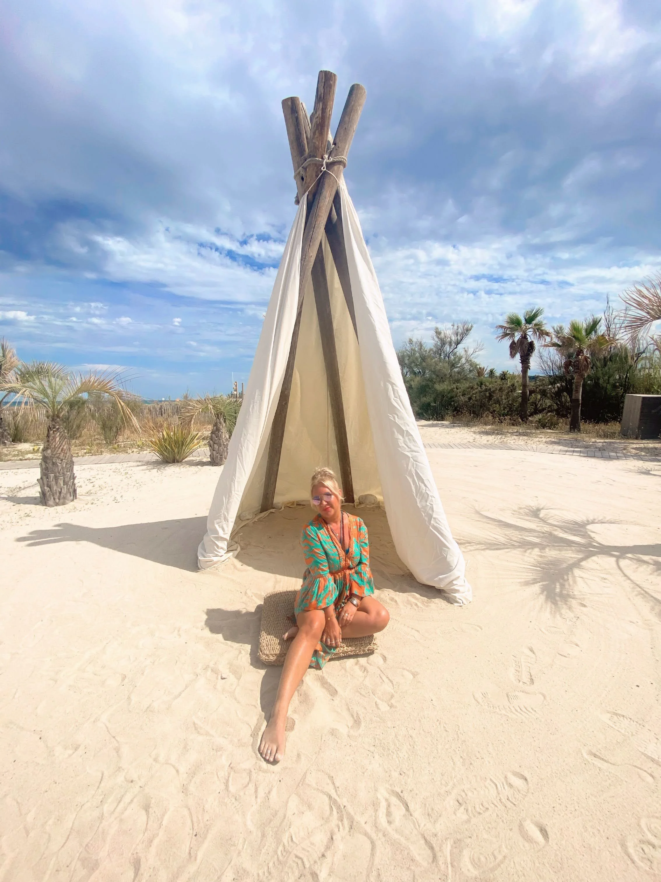 A woman sitting on a woven mat in front of a white fabric teepee on a sandy beach, with palm trees and a partly cloudy sky in the background.