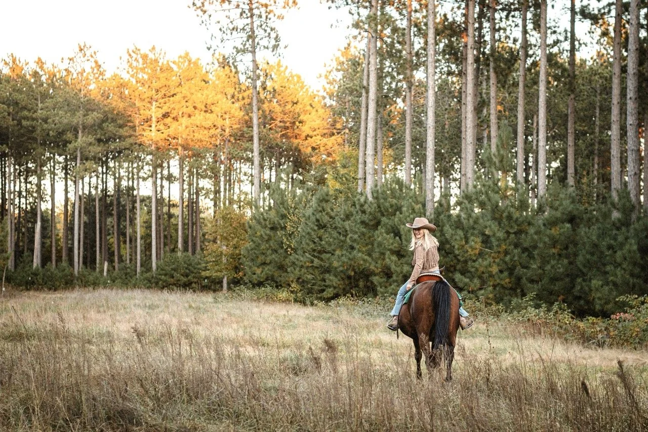 A woman riding a horse through a field with trees in the background during fall, wearing a cowboy hat and western attire.