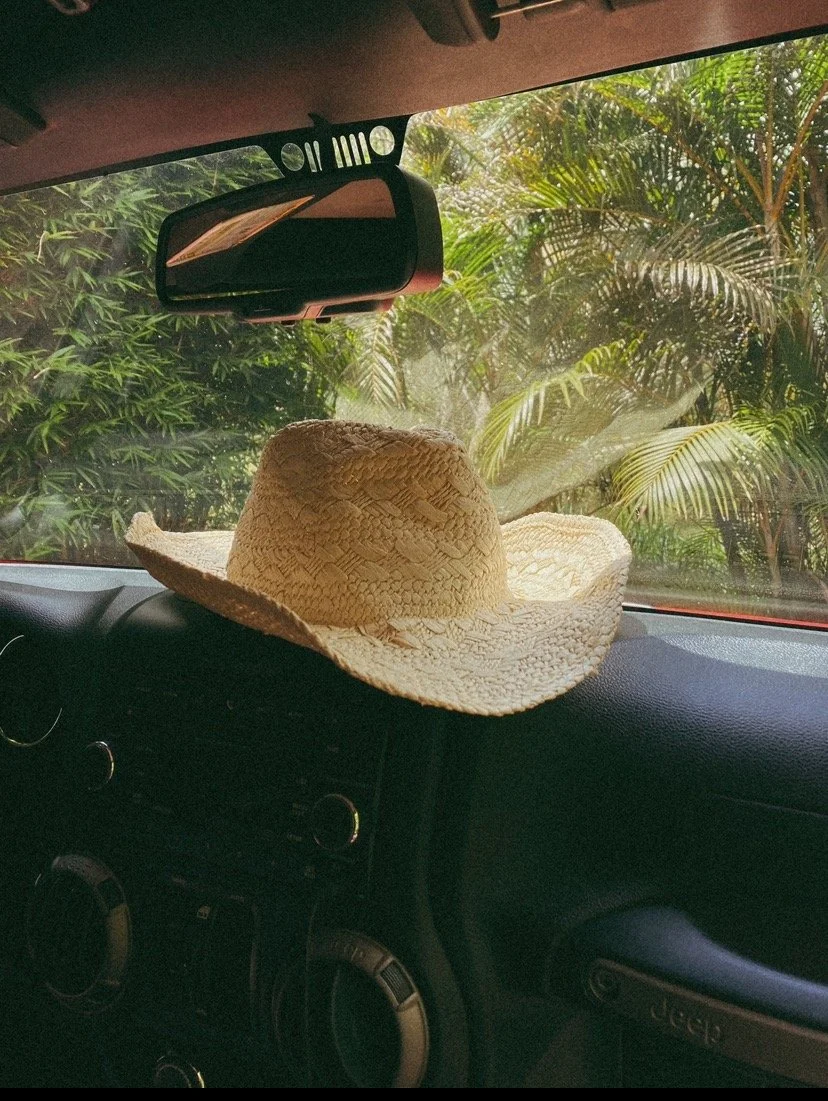 A straw cowboy hat resting on the dashboard of a Jeep vehicle, with tropical foliage visible outside the windshield.