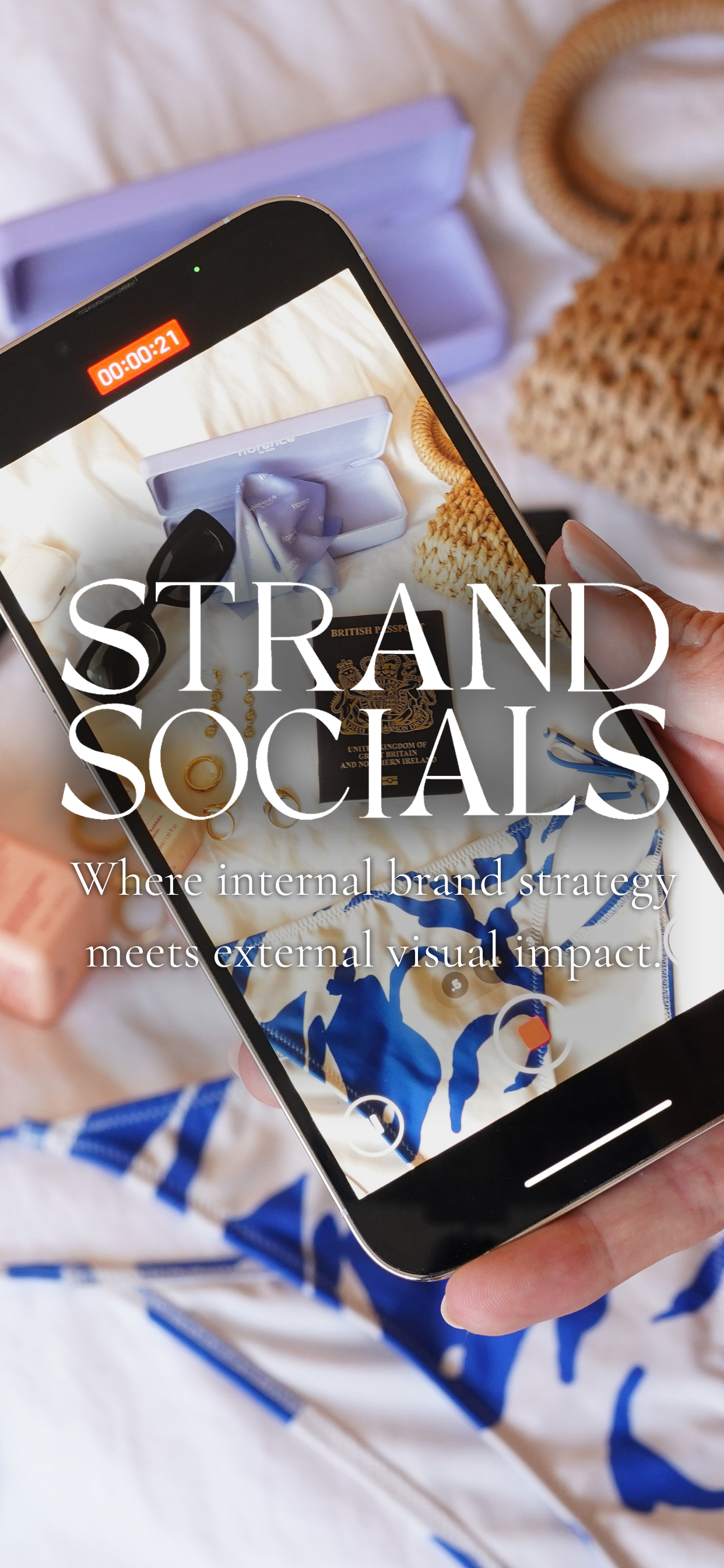 Smartphone displaying a photograph of various accessories including a British passport, gold rings, sunglasses, a lavender gift box, and a blue and white patterned item, on a bed with beige sheets, with a woven basket and purple box in the background.