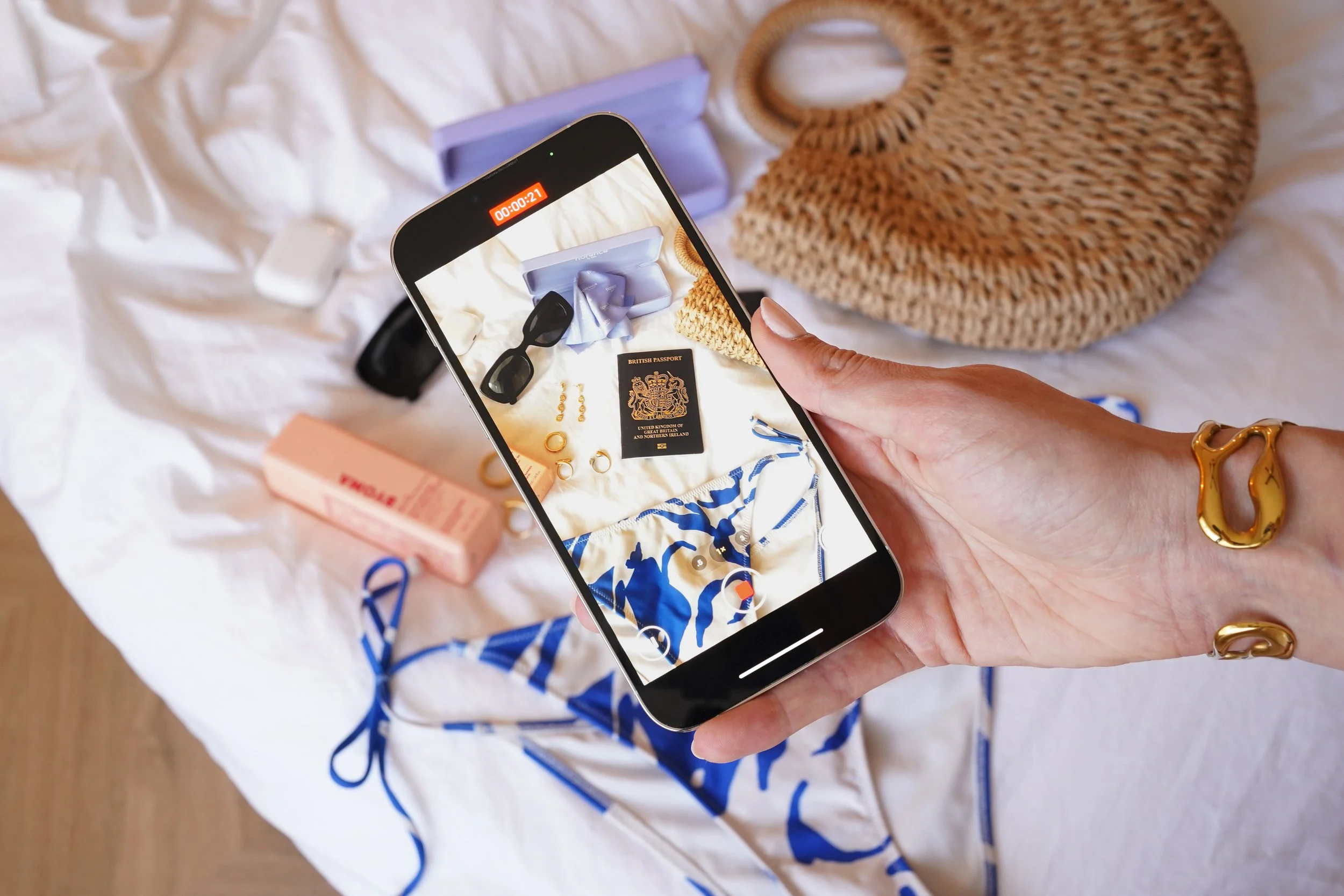 A person holds a smartphone taking a photo of various travel essentials laid out on a white bedsheet, including sunglasses, a British passport, jewelry, a makeup product, and a straw bag.