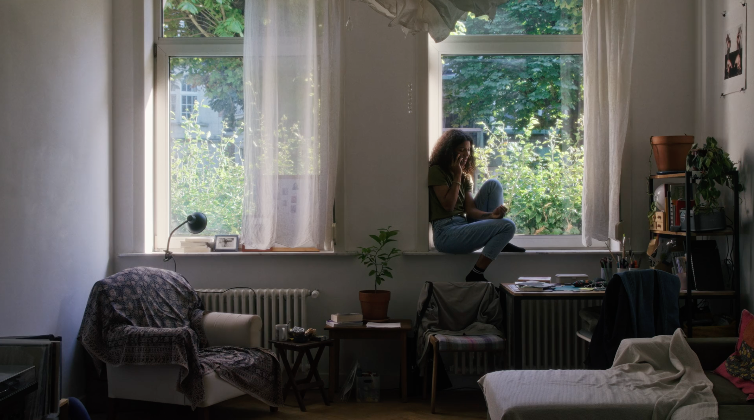 A woman sitting on a windowsill in a cluttered living room, talking on the phone. Behind her, large windows reveal green trees outside. The room has various furniture and plants, with sunlight streaming in.