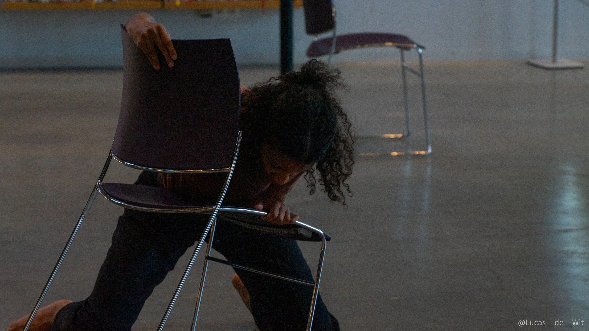 A woman with curly hair bowing deeply in front of a chair in a spacious room, with another empty chair in the background.