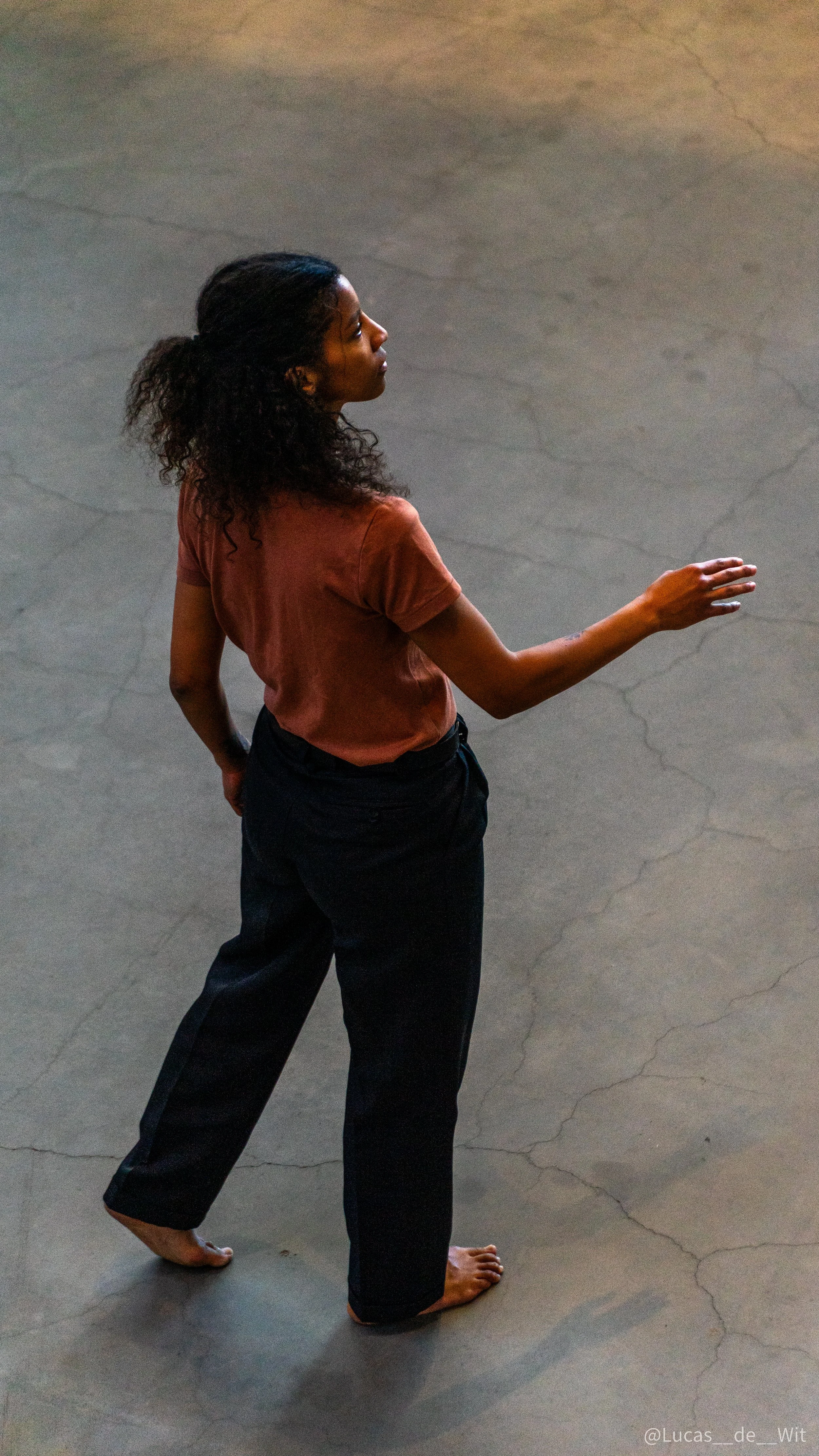 A woman with curly hair, wearing a brown t-shirt and black pants, standing barefoot on a concrete floor, gesturing with her right hand.