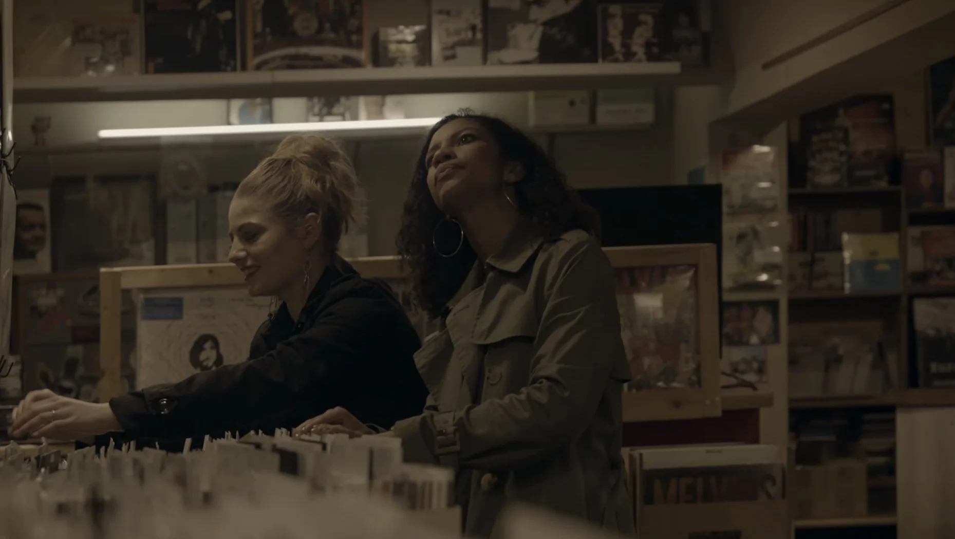 Two women shopping for vinyl records inside a record store, with shelves of records in the background.