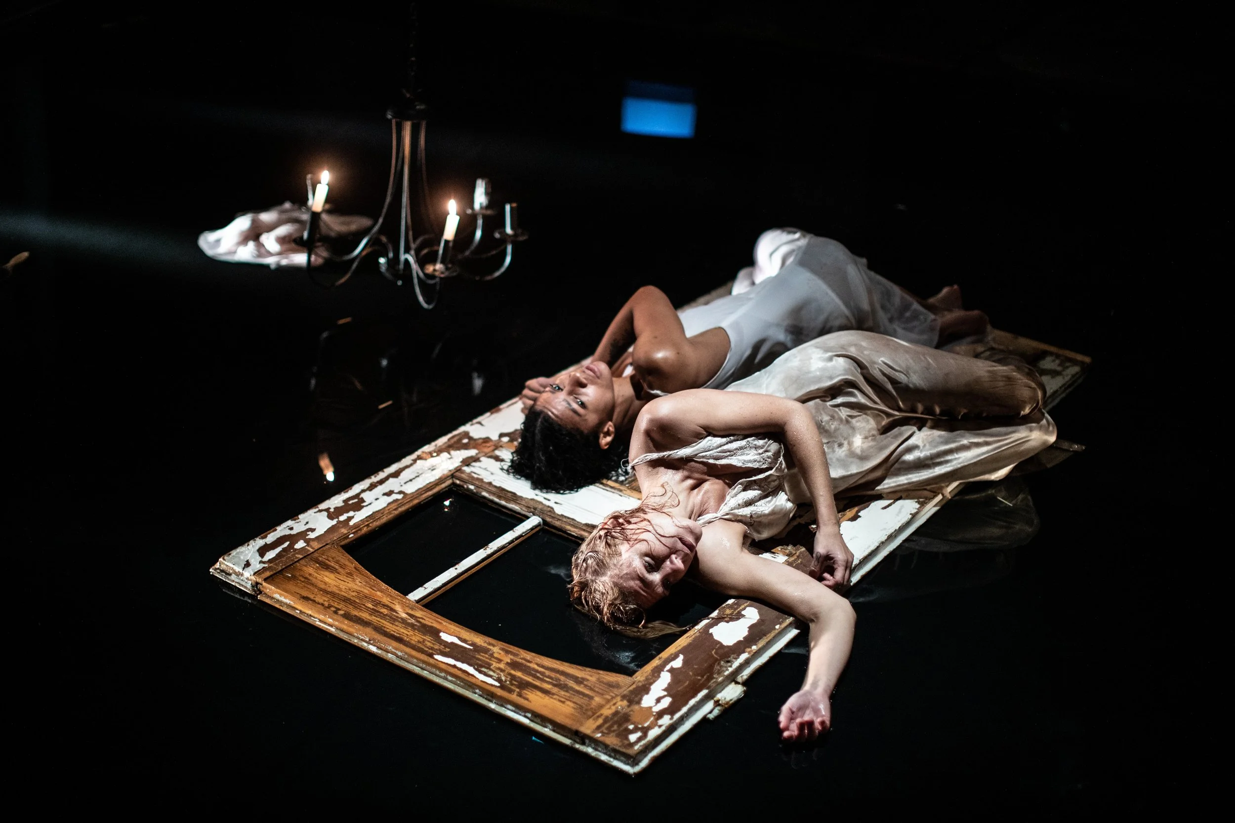 Two women lying on a worn wooden frame on a dark floor, with a chandelier hanging above them