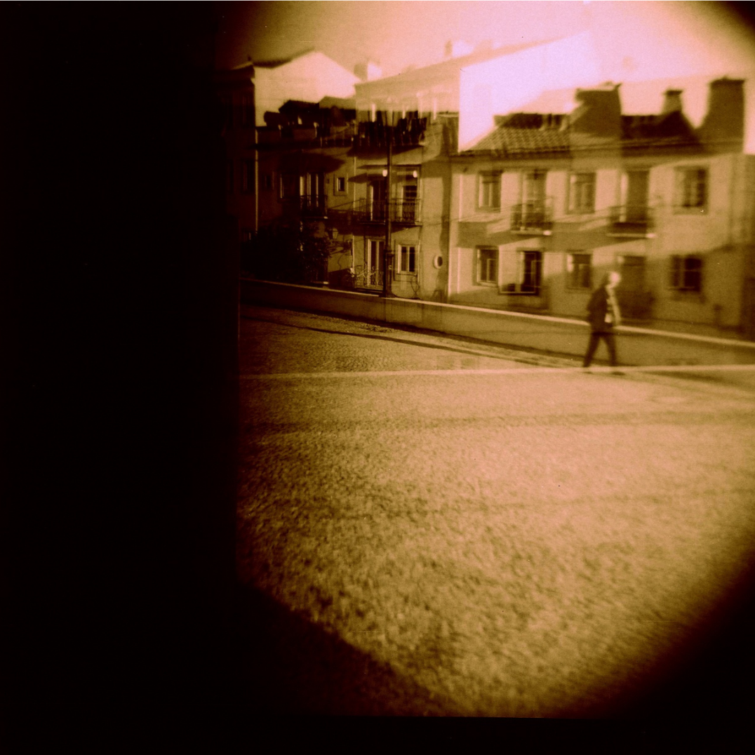 A sepia-toned photo of a man walking on a city street with residential buildings in the background, viewed through a circular lens or peephole.