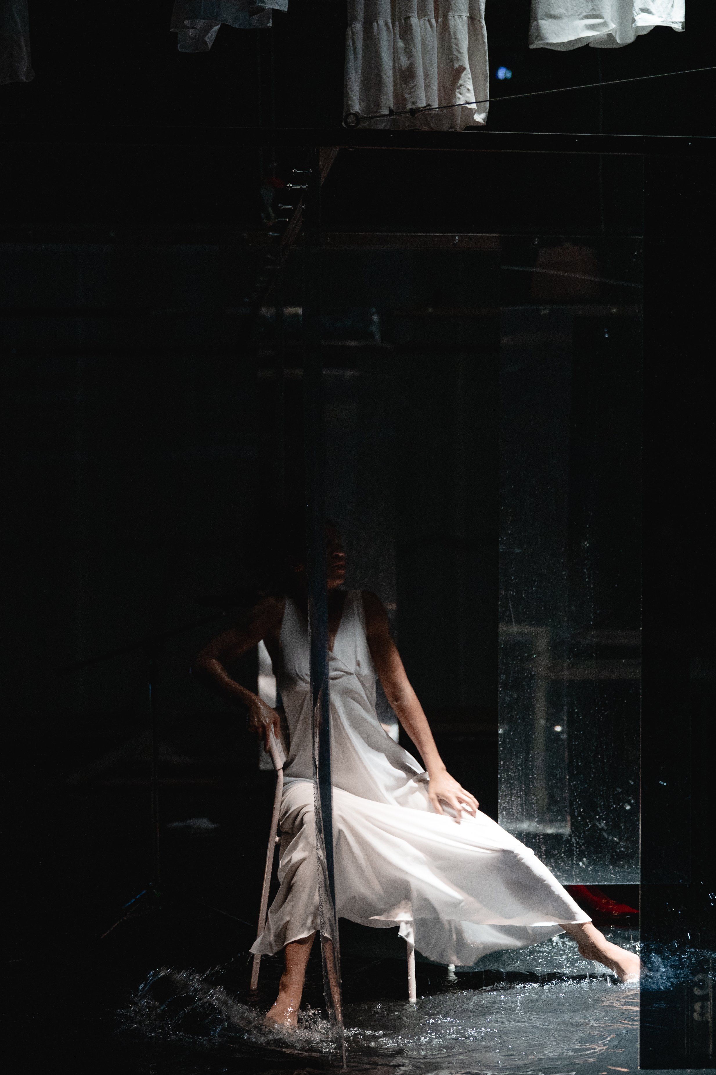 A woman in a white dress sitting on a chair with her feet in water, inside a dark room with mirrors and laundry hanging above.