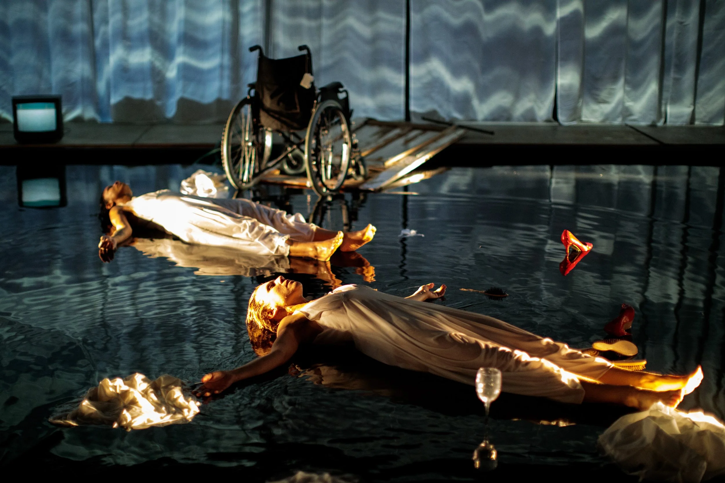 Two women in white dresses lying in a body of water, with the reflection of the women and surrounding objects visible on the water's surface. A wheelchair and a small television are positioned on a platform in the background, and a broken high-heeled