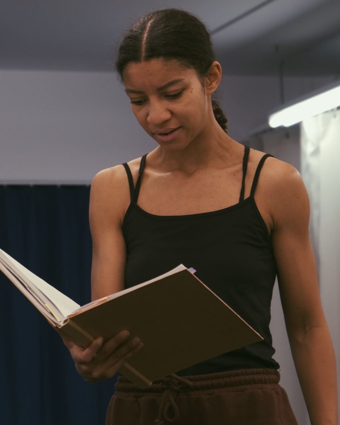 A woman in a black tank top and brown sweatpants reading a large book in a room with soft lighting.