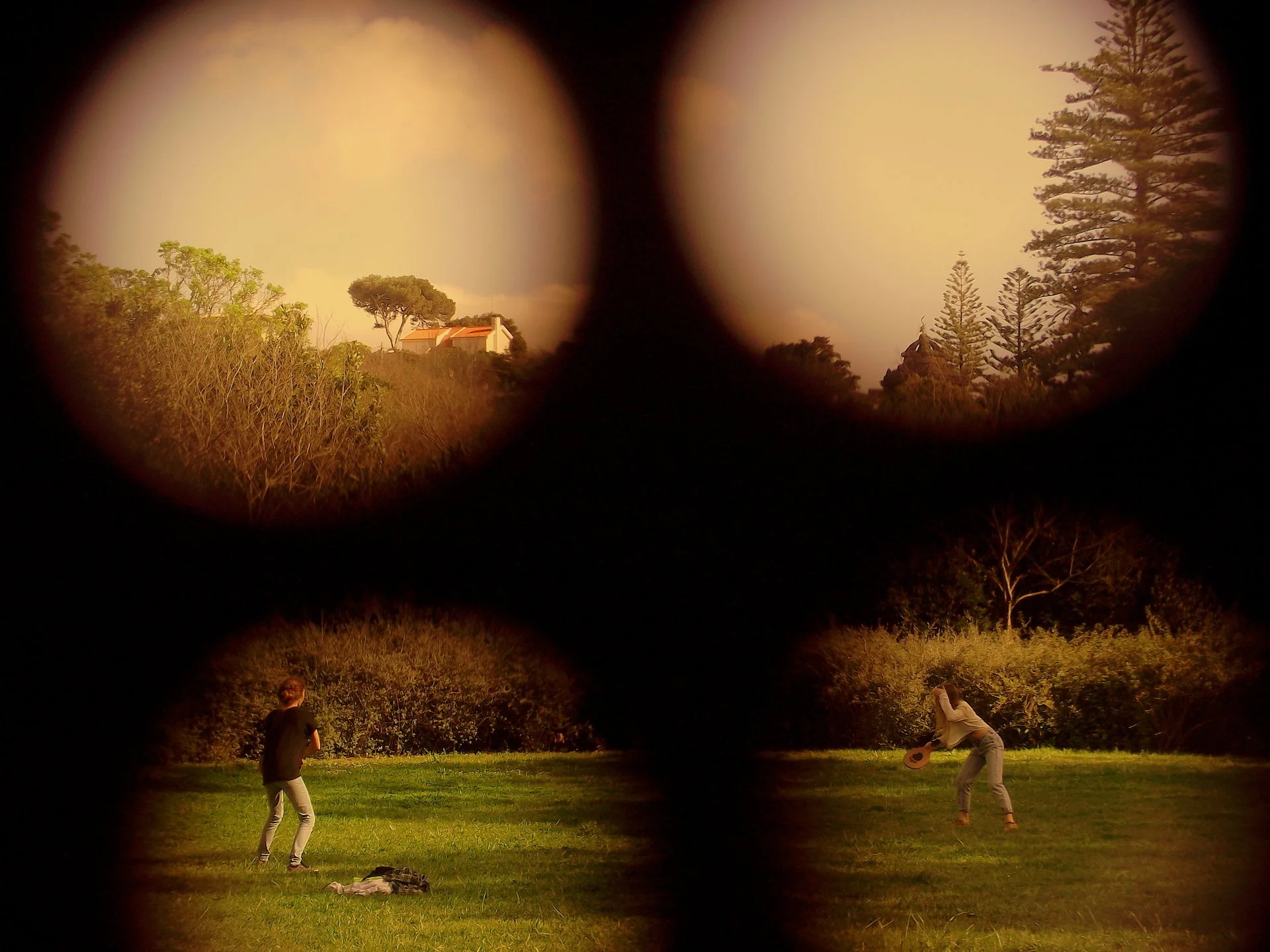 The image shows two people playing frisbee on a grassy field, viewed through binoculars. In the background, there are trees and a house with a red roof under a partly cloudy sky.
