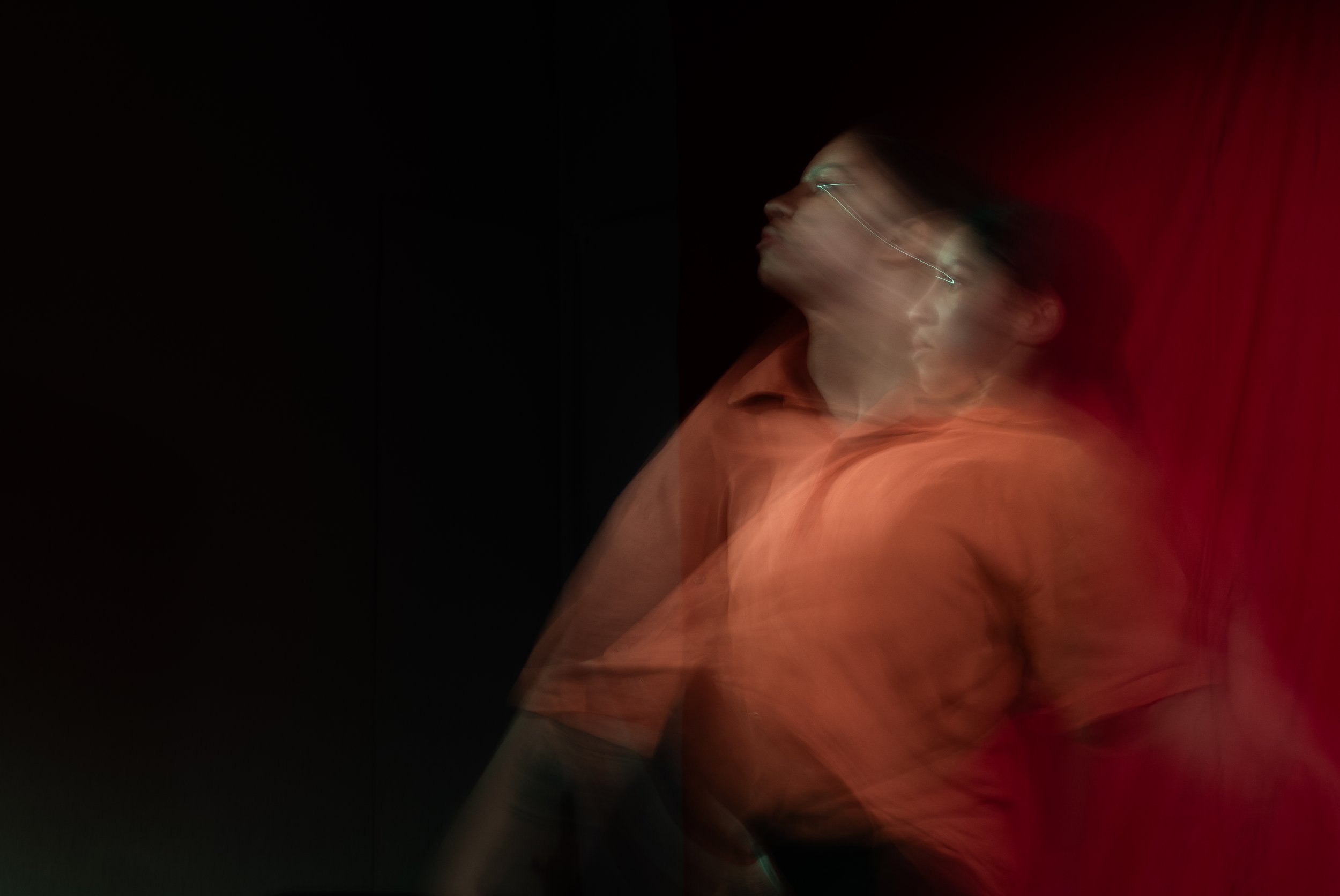 A person with glasses in a motion blur, wearing an orange top, with a dark background and a red curtain on the right.