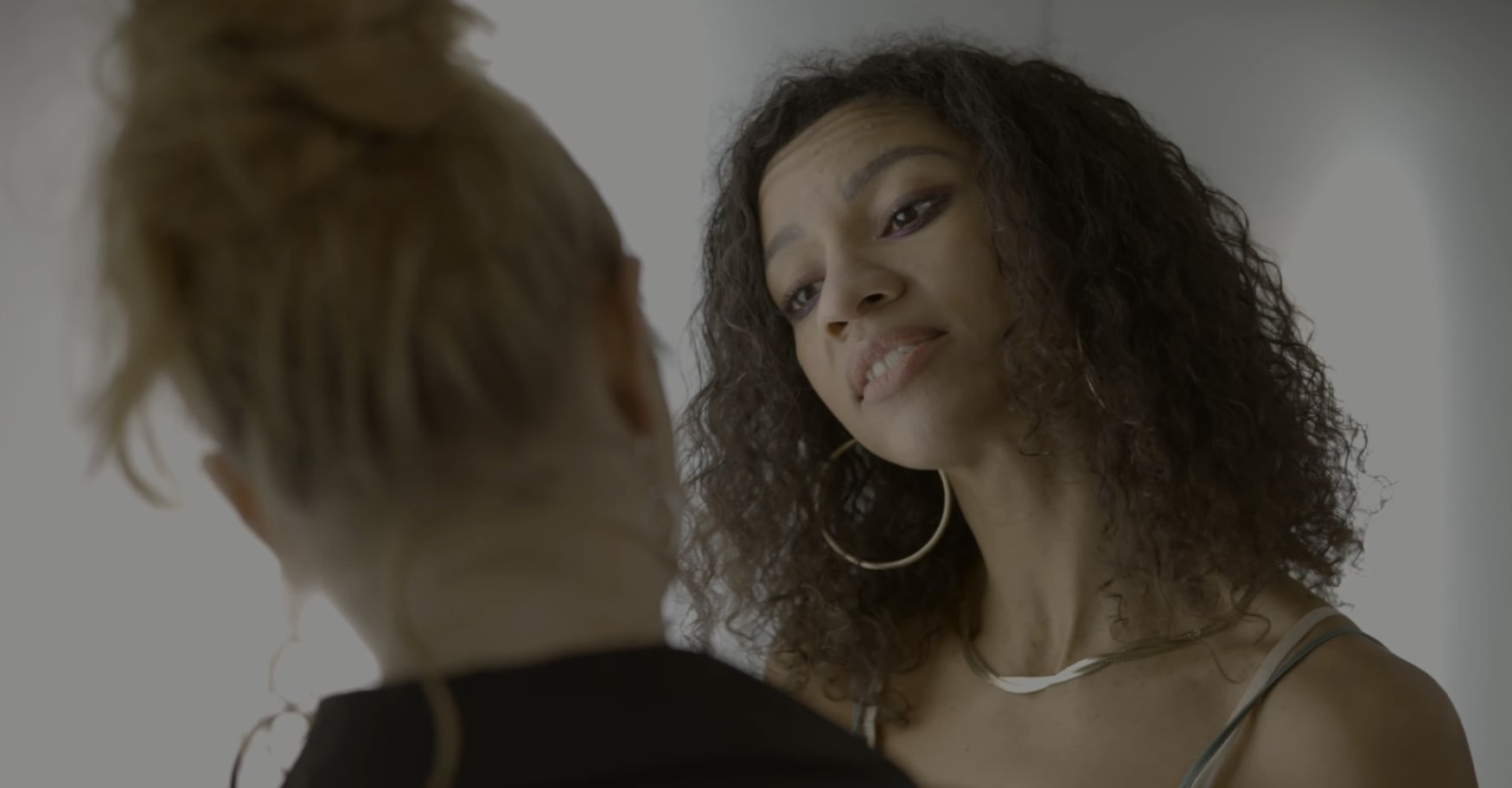 Two women facing each other, one with curly hair and hoop earrings, smiling gently.