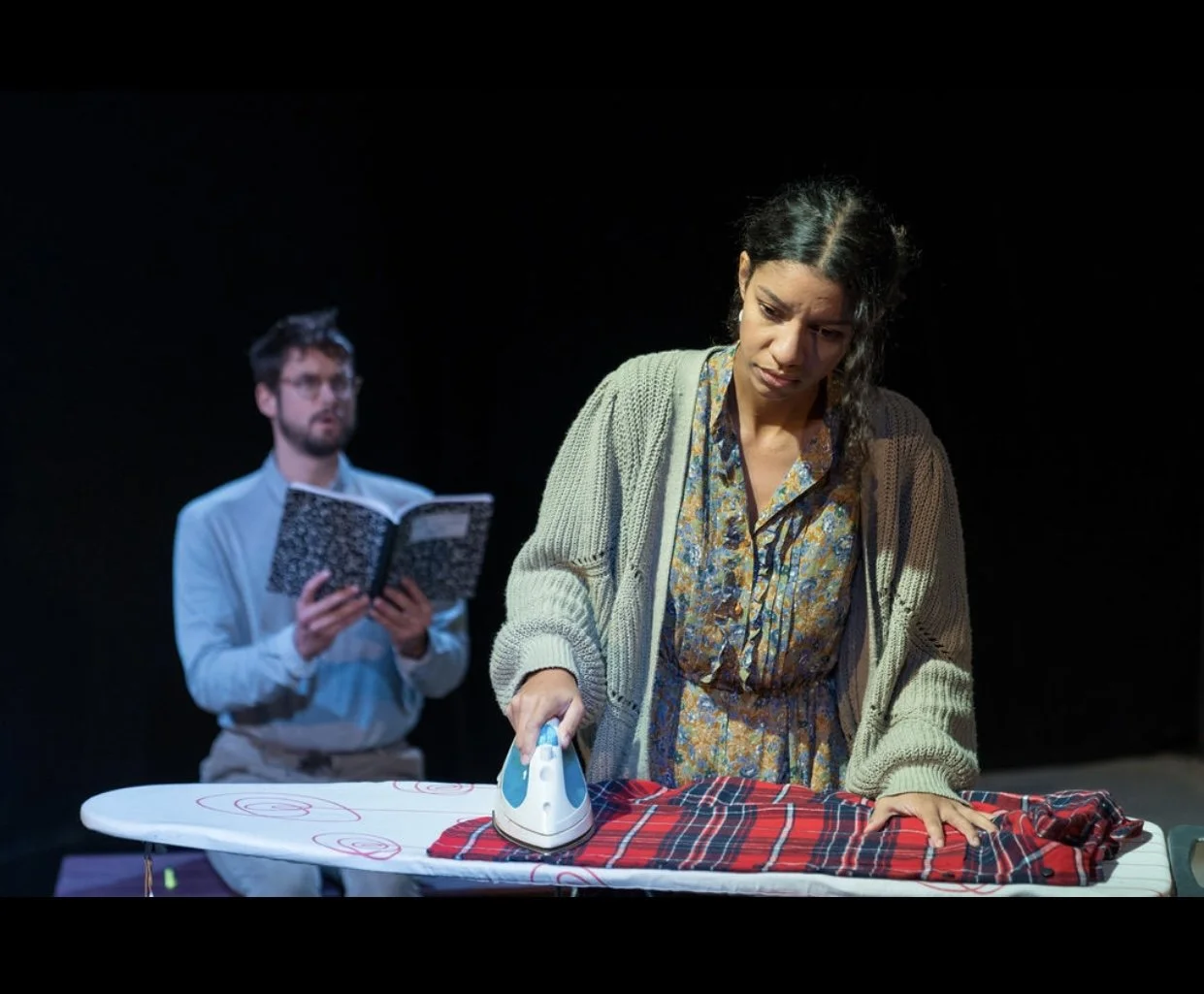 A woman ironing a piece of red and black plaid fabric on an ironing board, with a man in the background reading a book.