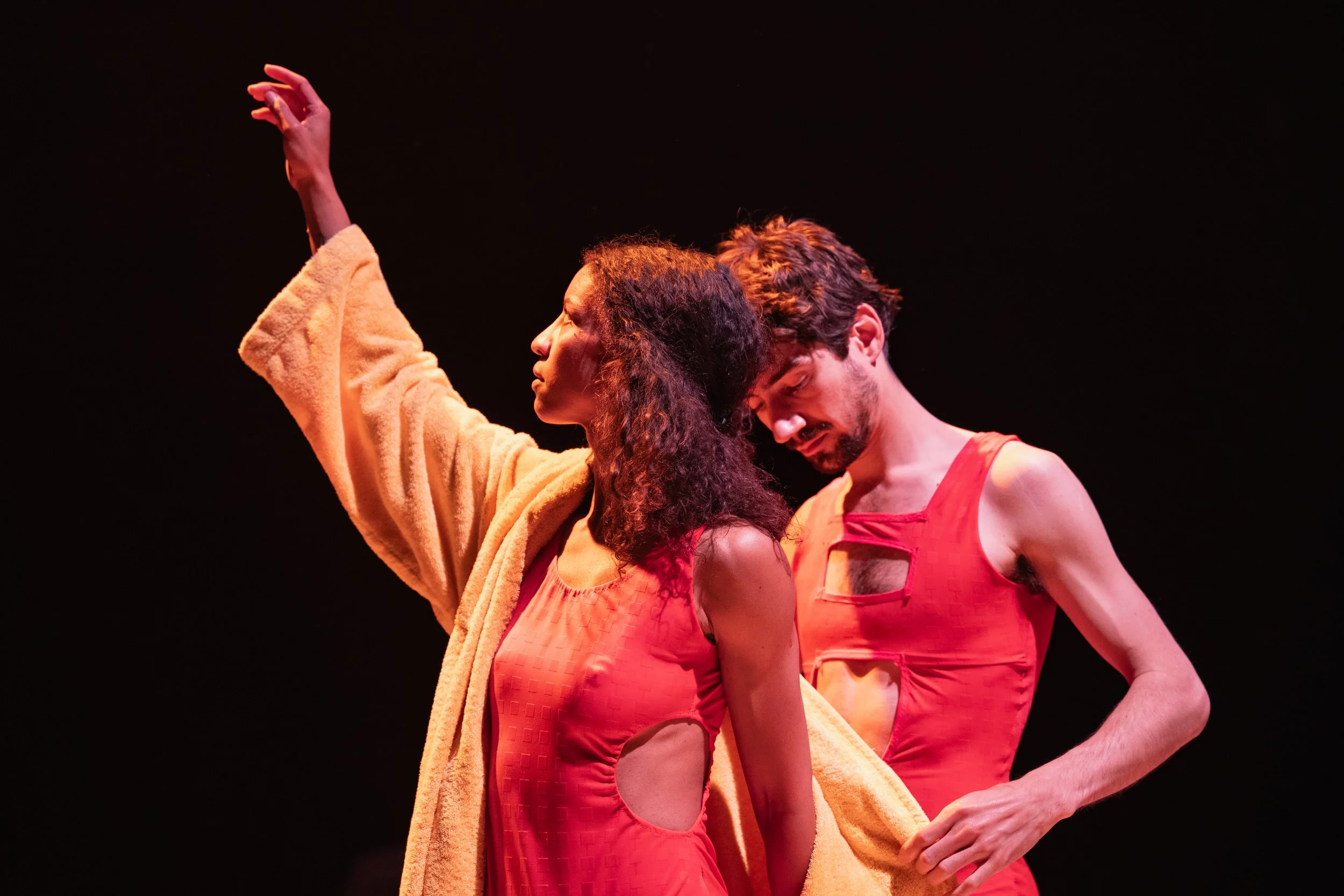 Two performers on stage, one with long curly hair and the other with short hair, wearing red outfits with cutouts, under warm lighting, against a dark background.