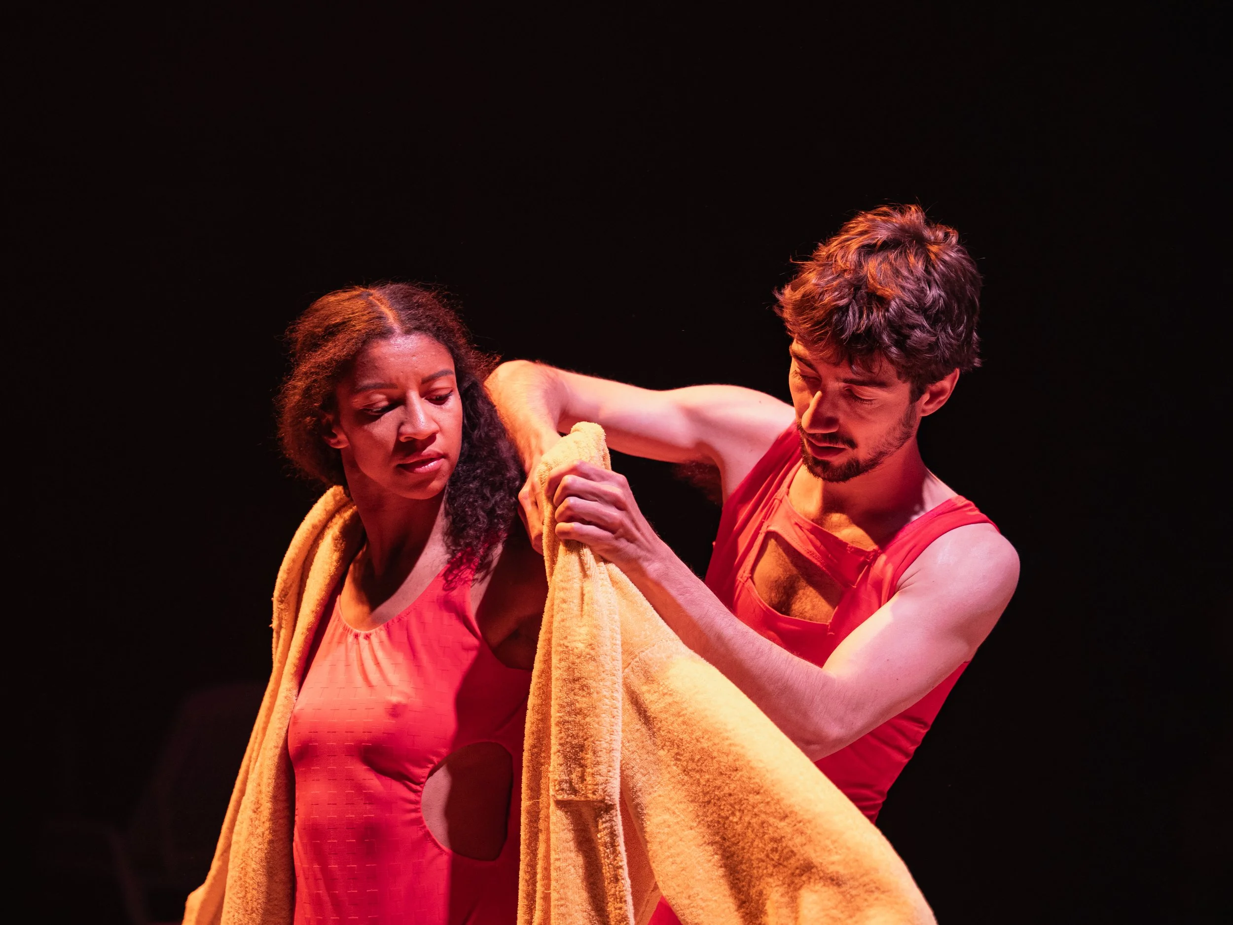 A man helping a woman remove a towel in a dance performance on stage with black background.