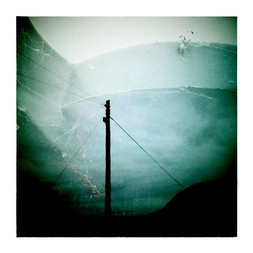 Photograph of a utility pole with power lines against a cloudy sky, with a cracked glass overlay and spider webs.