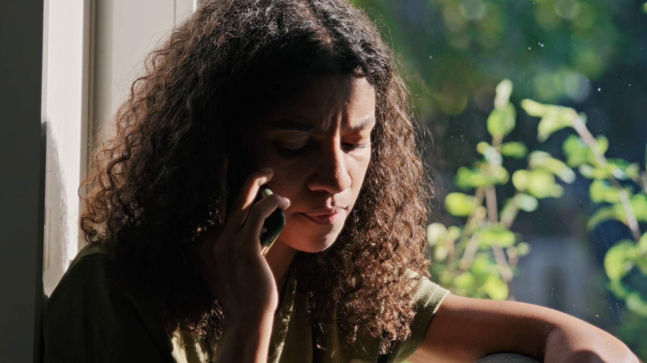 A young woman with curly hair looking concerned, talking on her cell phone, sitting by a window with sunlight and greenery visible outside.