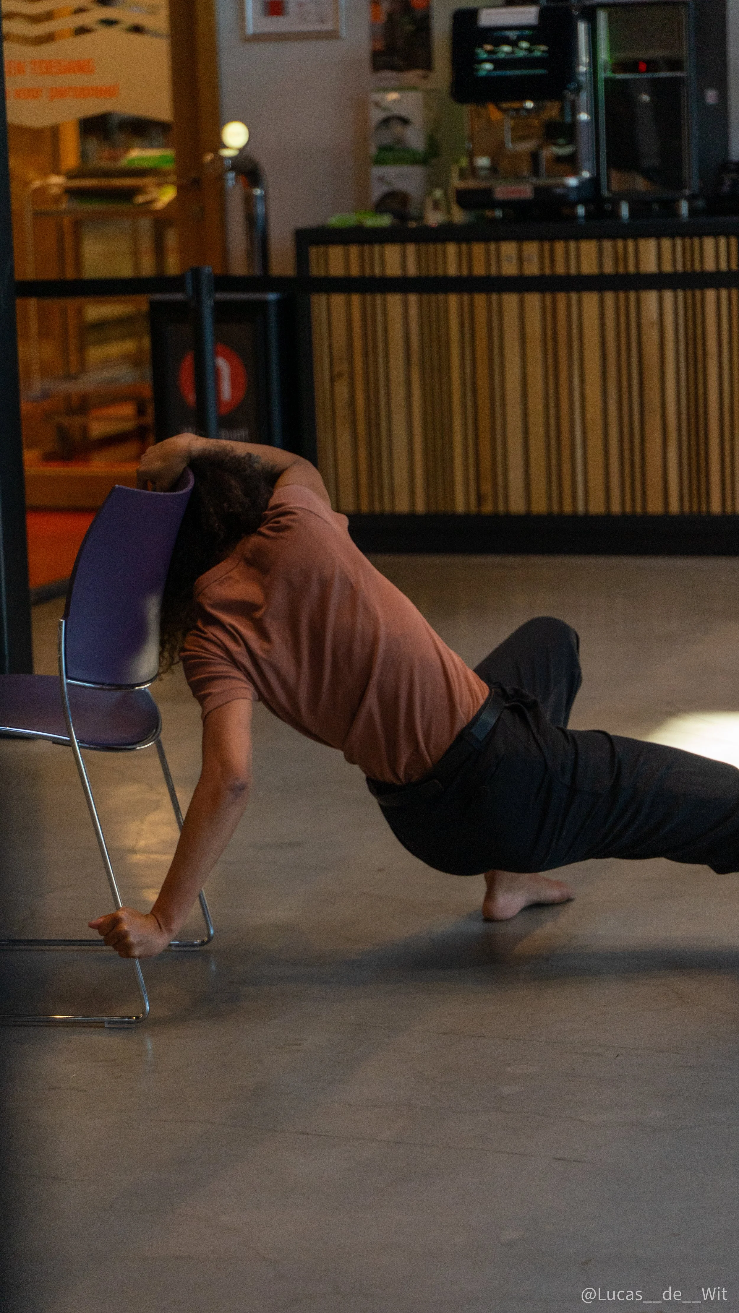 A person with curly hair performing a floor exercise in a cafe or restaurant setting, supporting themselves with one hand on the floor and the other on a chair, with a coffee shop background.