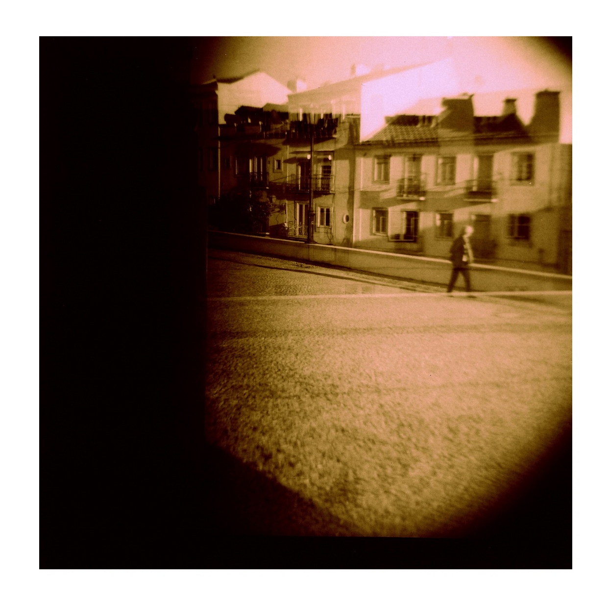 A person walking on a city sidewalk with residential buildings in the background, seen through a distorted, sepia-toned or underwater-like lens effect.