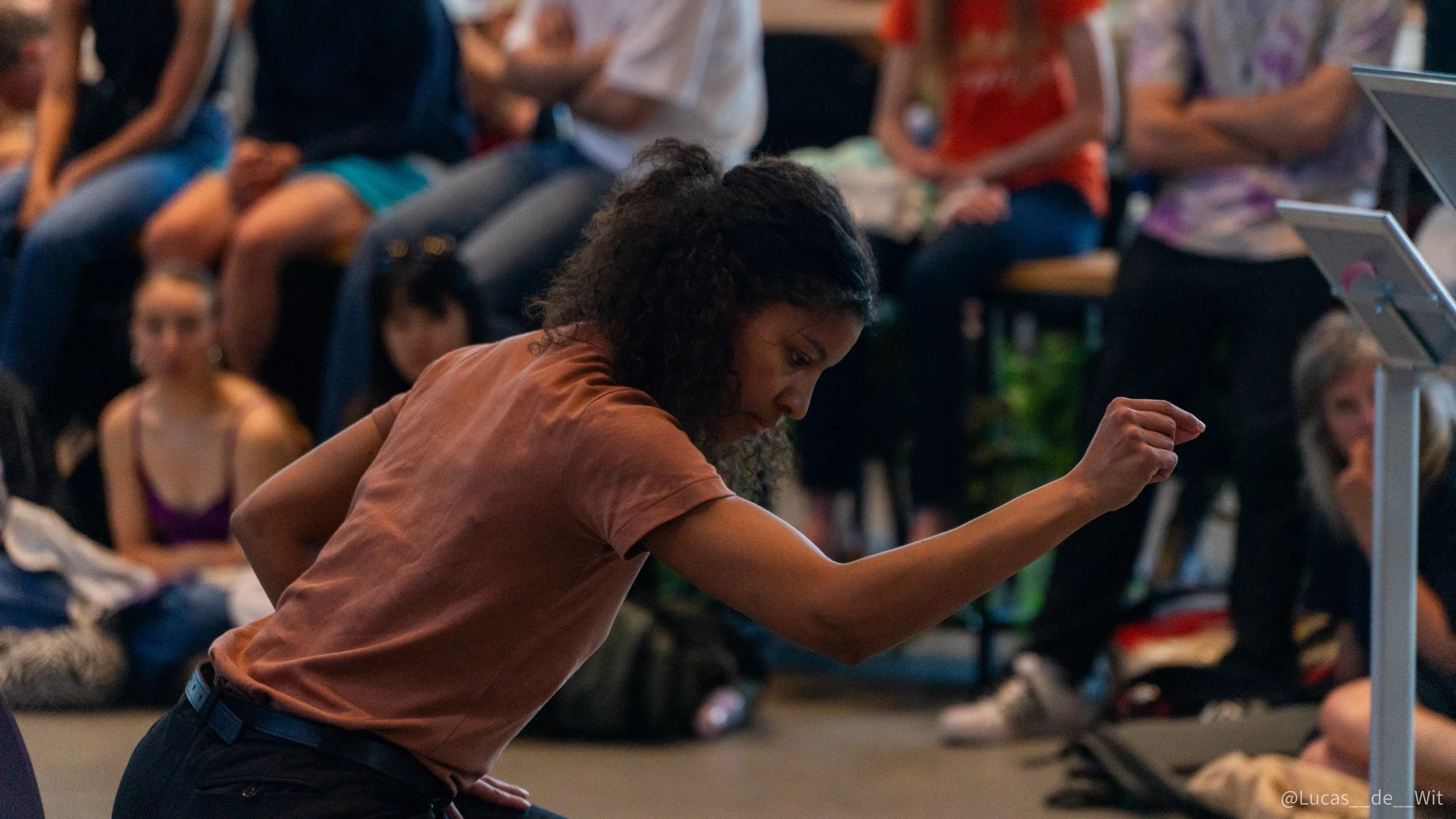A woman with curly hair wearing a brown T-shirt leaning forward and reaching out with her right hand in a crowded indoor setting with seated people in the background.