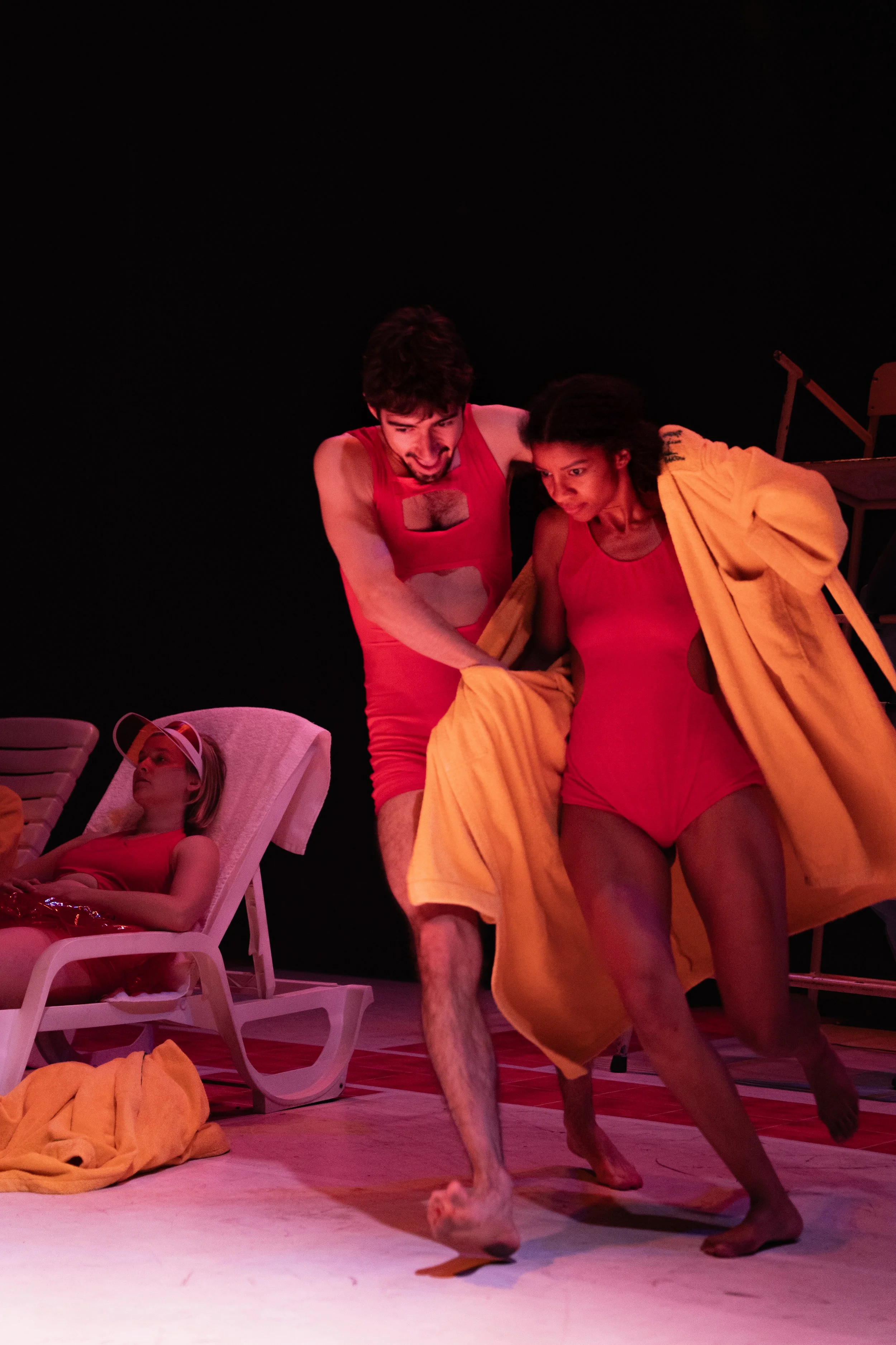 Three performers on stage dressed in red and yellow costumes, with one lying on a lounge chair in the background and two others engaged in a scene with lively expressions.