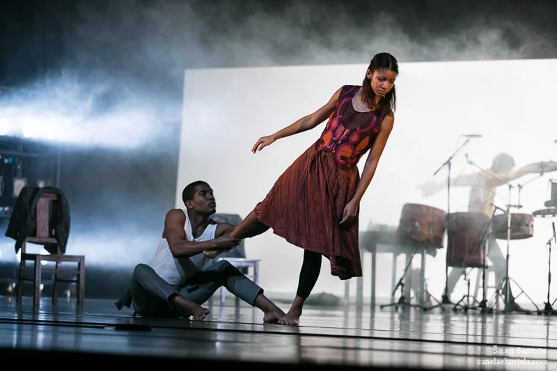 Two dancers performing on stage with a band in the background, one sitting on the floor and holding the ankle of the other, who is standing and leaning forward.