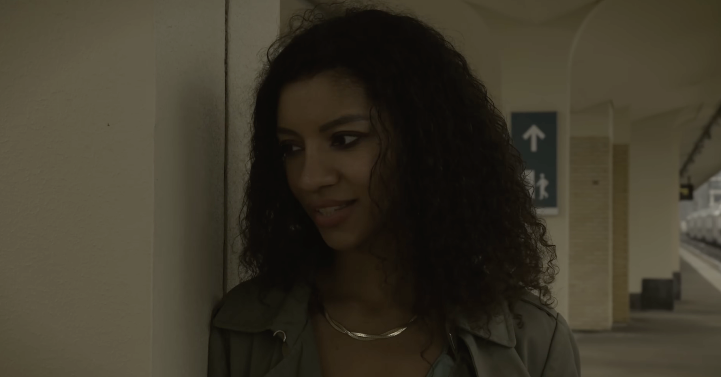 A woman with curly hair standing against a wall in a train station, looking to her left.