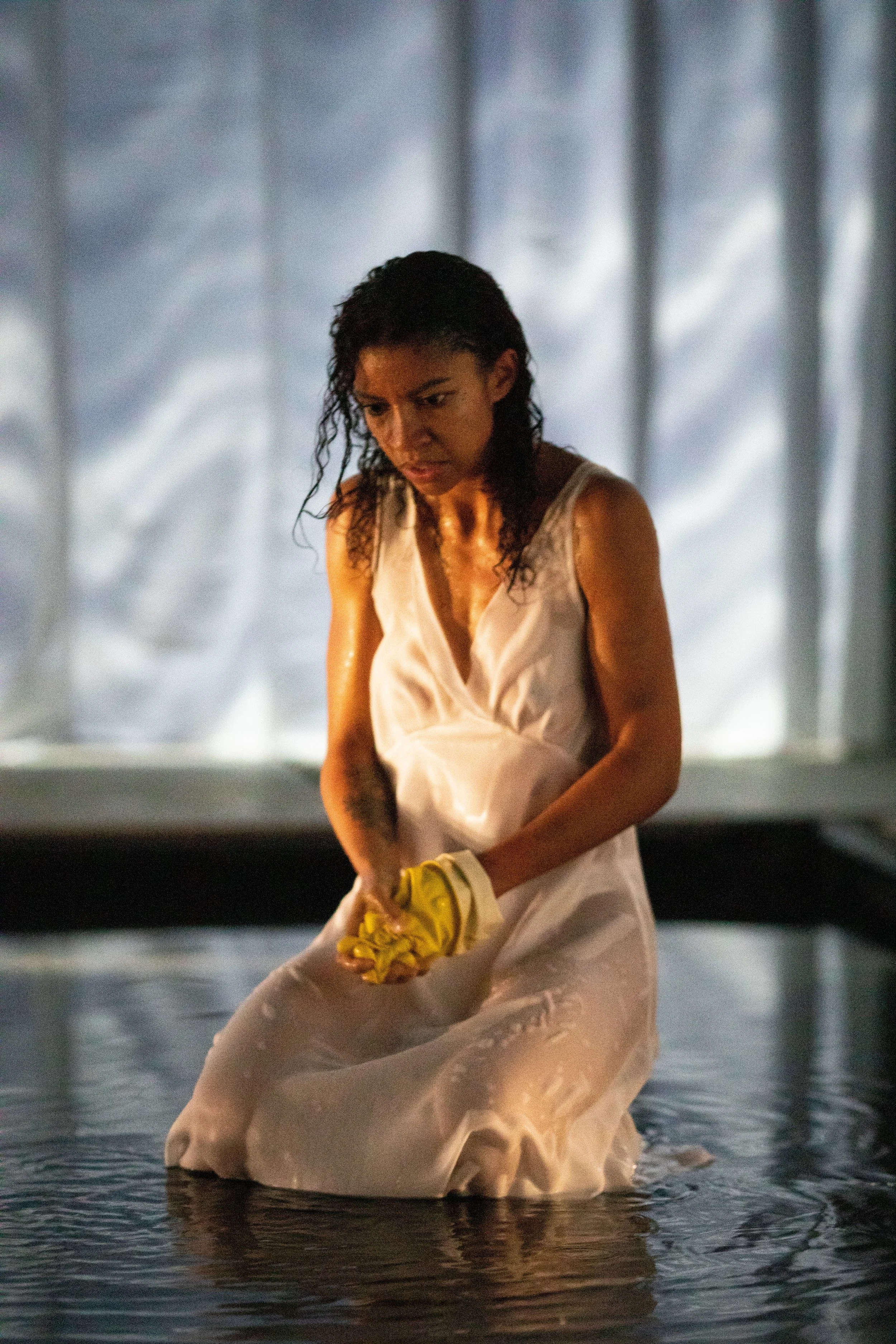 Woman in a white dress kneeling in water, holding a yellow cloth, with a sad or distressed expression.