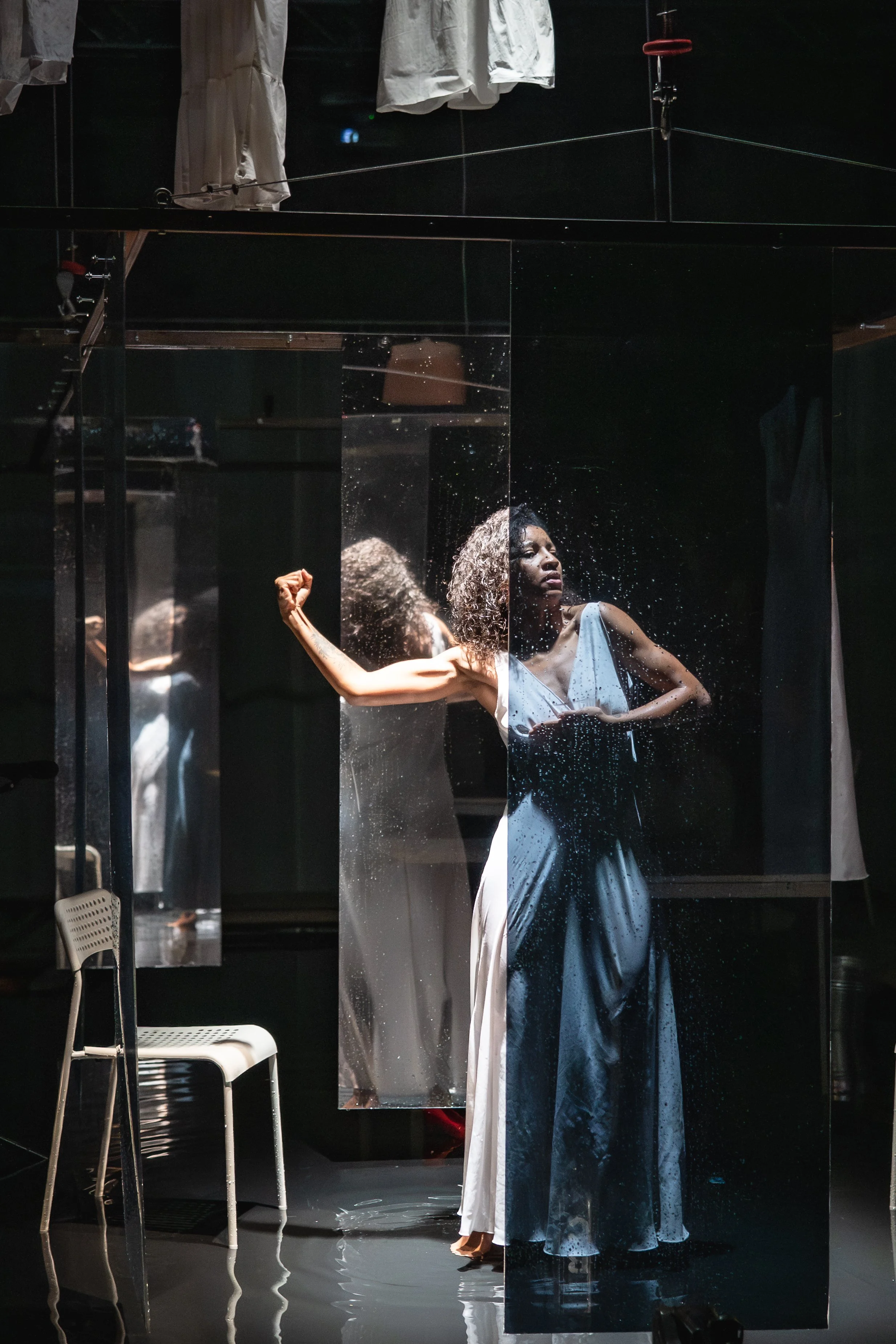 A woman with curly hair is standing in front of mirror panels, flexing her arm muscles while wearing a sleeveless dress. The reflection shows her profile, and there is water on the mirrors.