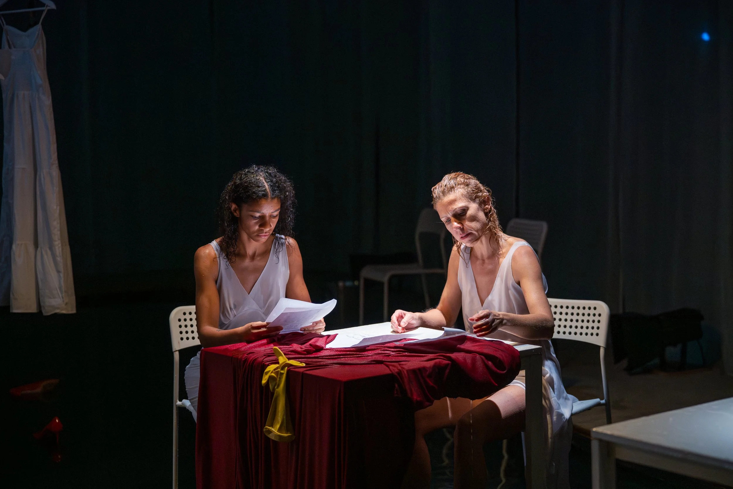 Two women sitting at a table with red fabric, looking at papers in a dimly lit room, possibly rehearsing or reading scripts.