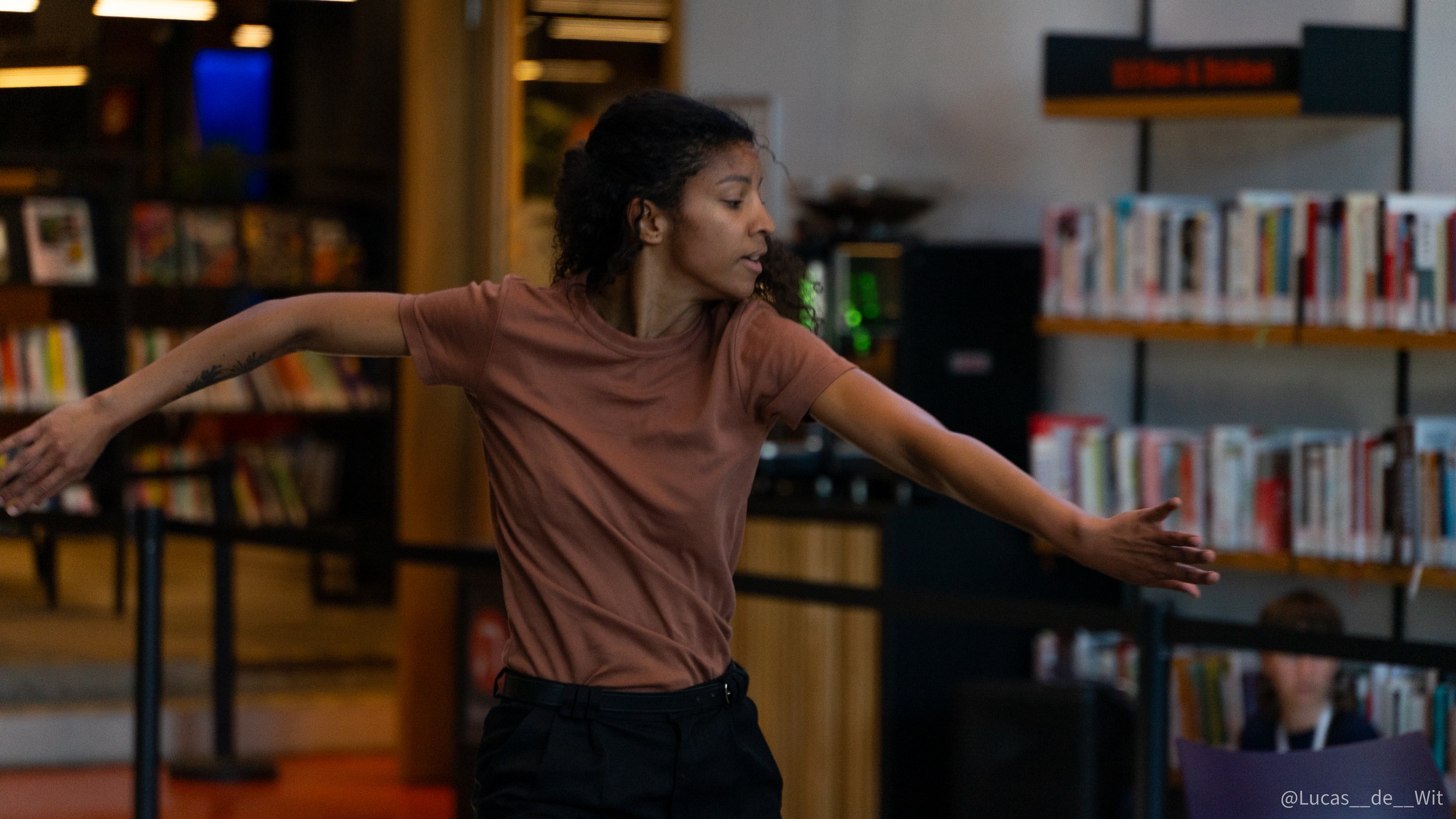 A woman with curly hair dancing or moving her arms in a library or bookstore setting, surrounded by bookshelves filled with books.