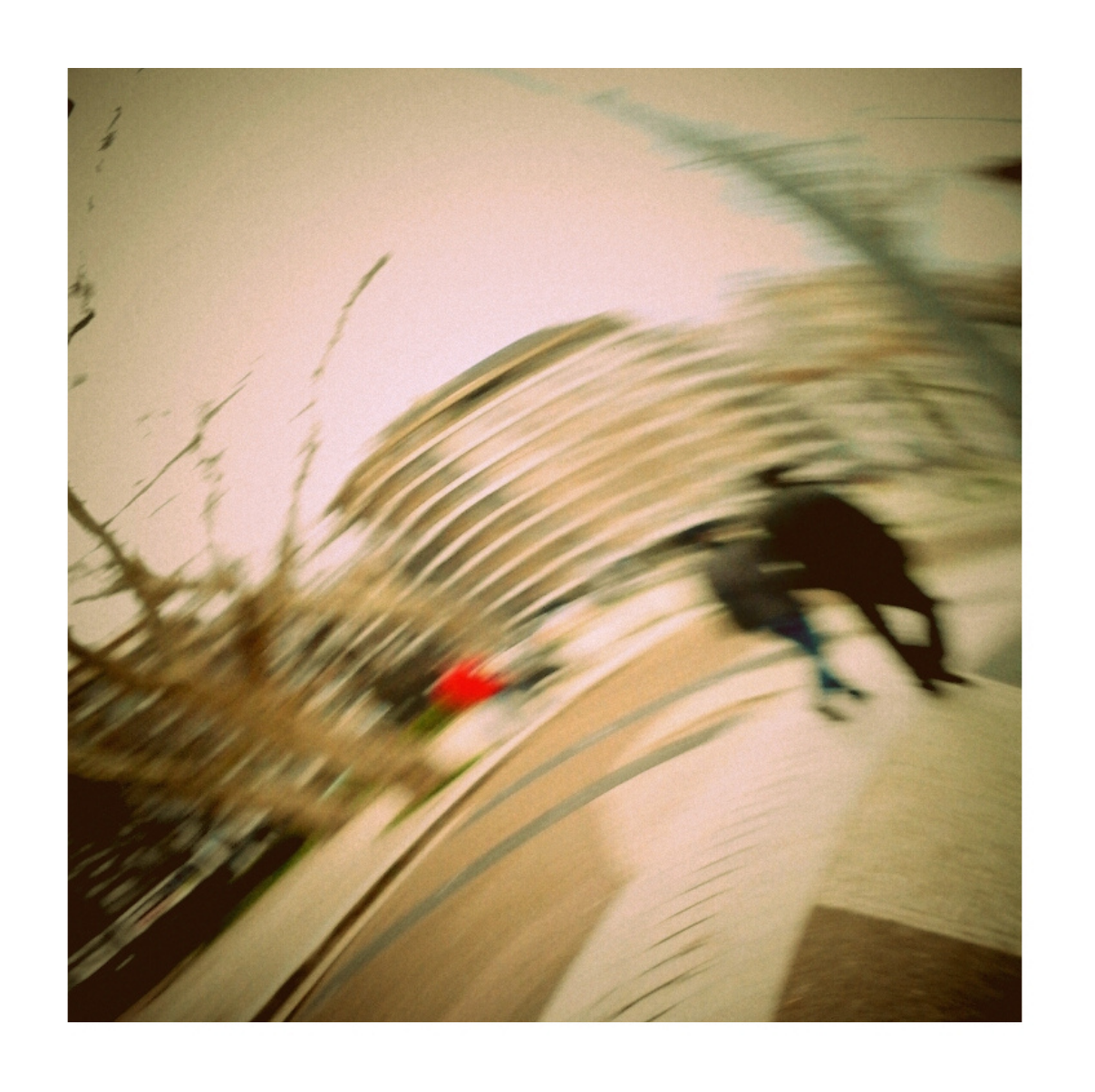 Blurred image of people walking outside near a curved roof or awning with tree branches and a red object in the foreground.