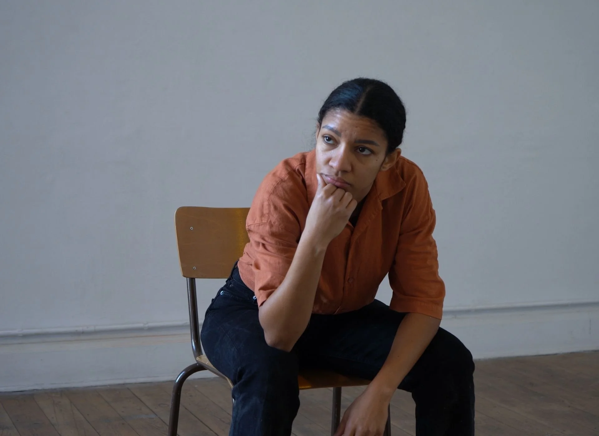 A woman with dark hair in a bun, wearing an orange shirt and black pants, sitting on a wooden chair with a thoughtful expression, resting her chin on her hand, in a room with a plain light-colored wall.