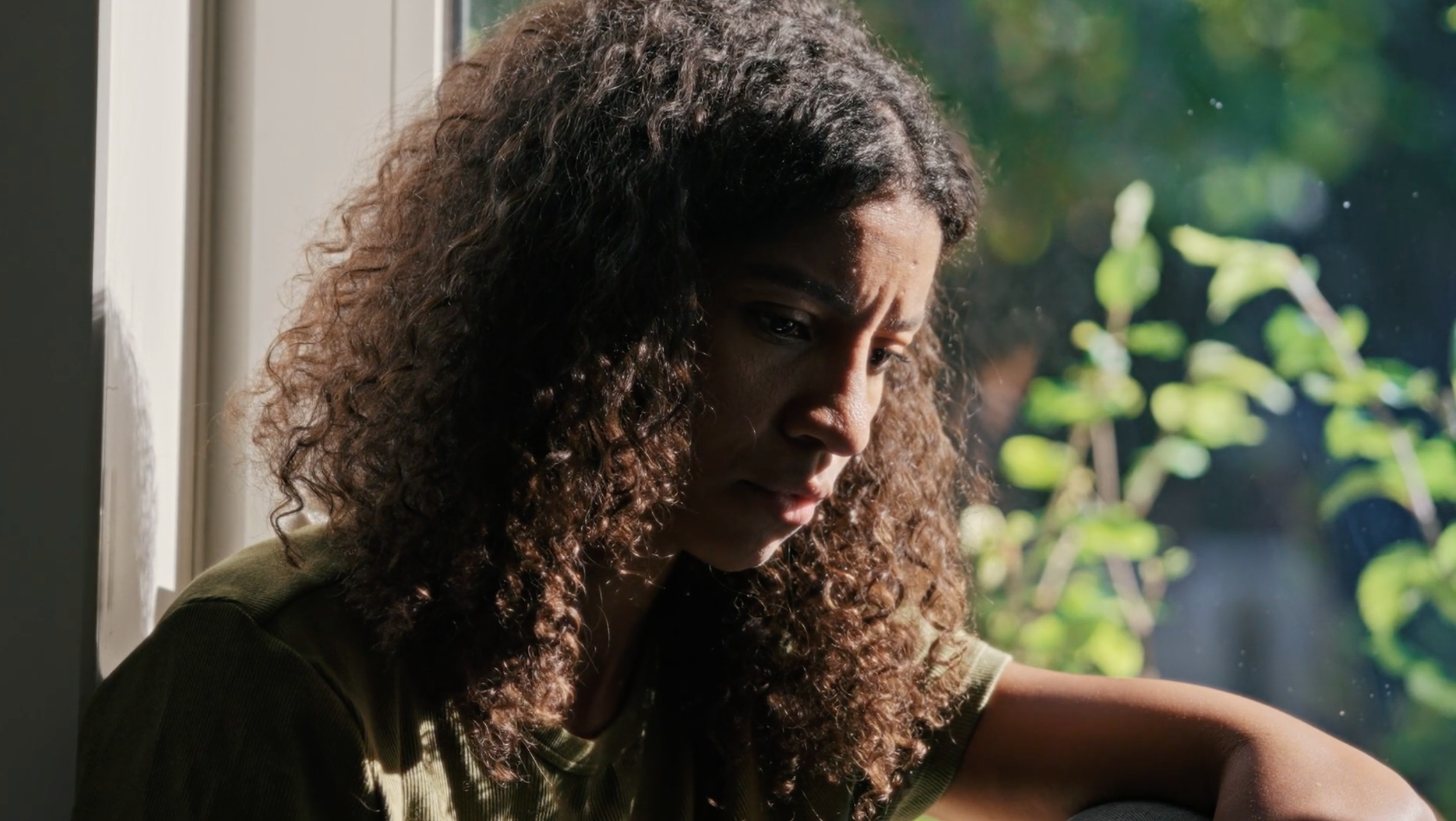 A woman with curly hair looking thoughtfully by a window with sunlight and greenery outside.