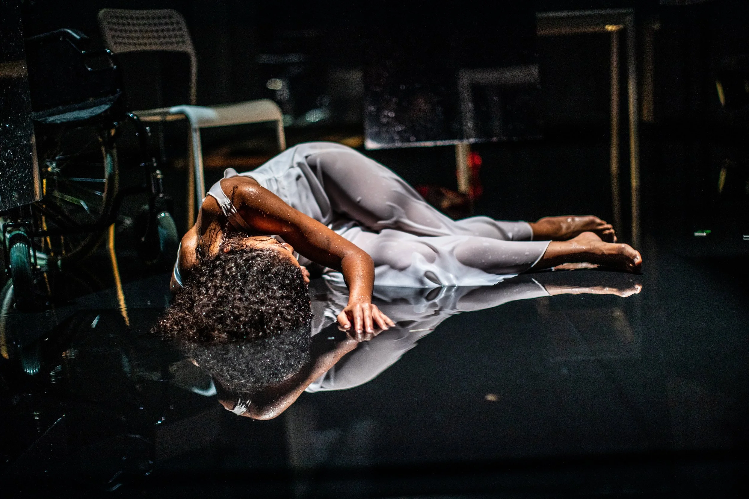 A woman lies on a shiny black floor, wearing a white dress, with her head turned to the side and her curly hair spread out, surrounded by scattered pool water and wetness.
