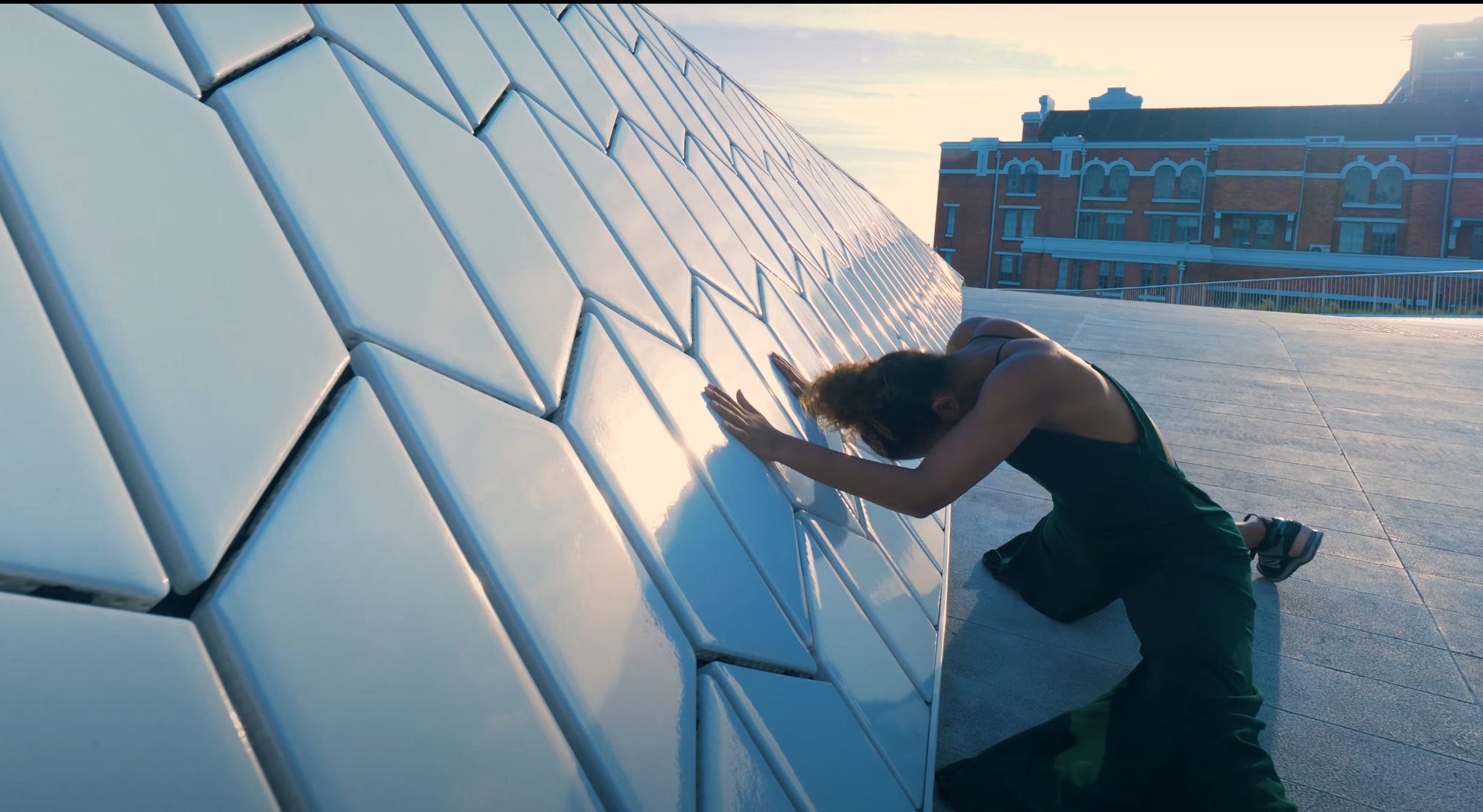 A woman with curly hair, wearing a black tank top and dark green pants, kneeling and touching a large, reflective, geometric white tiled wall outdoors at sunset.