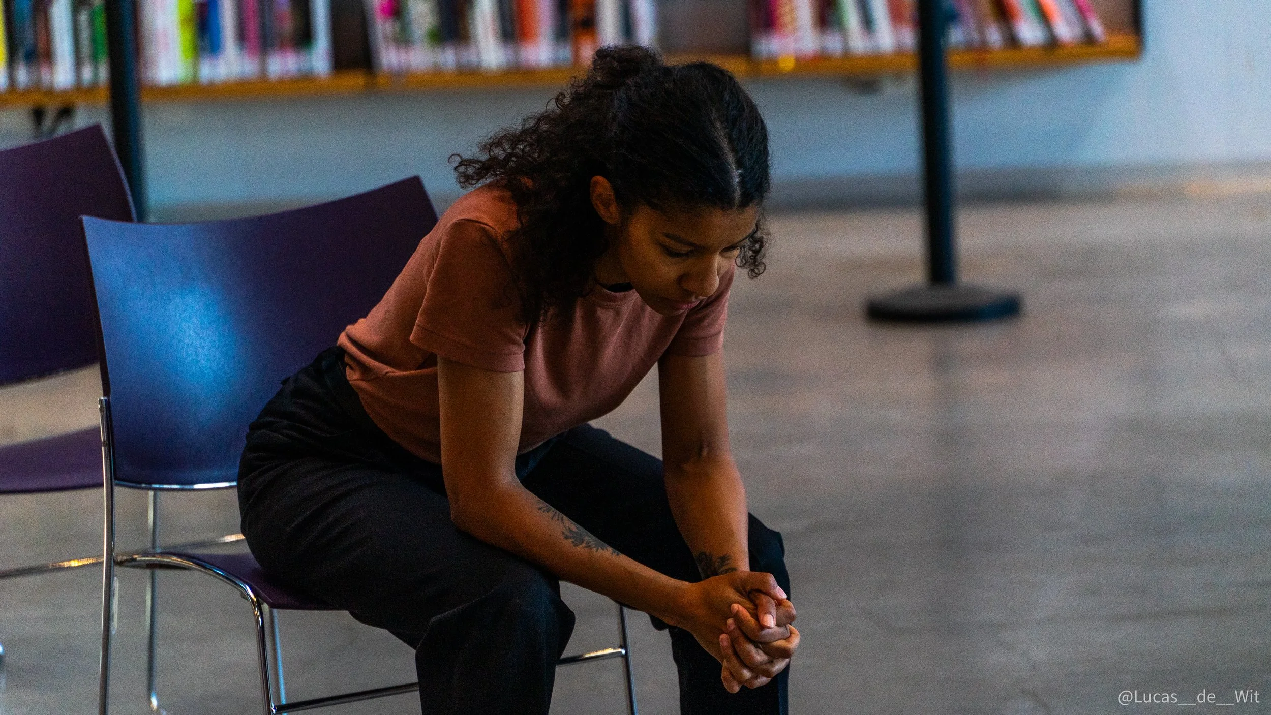 A woman with curly dark hair is sitting on a chair with her head bowed and hands clasped in front of her, in a library or similar setting.