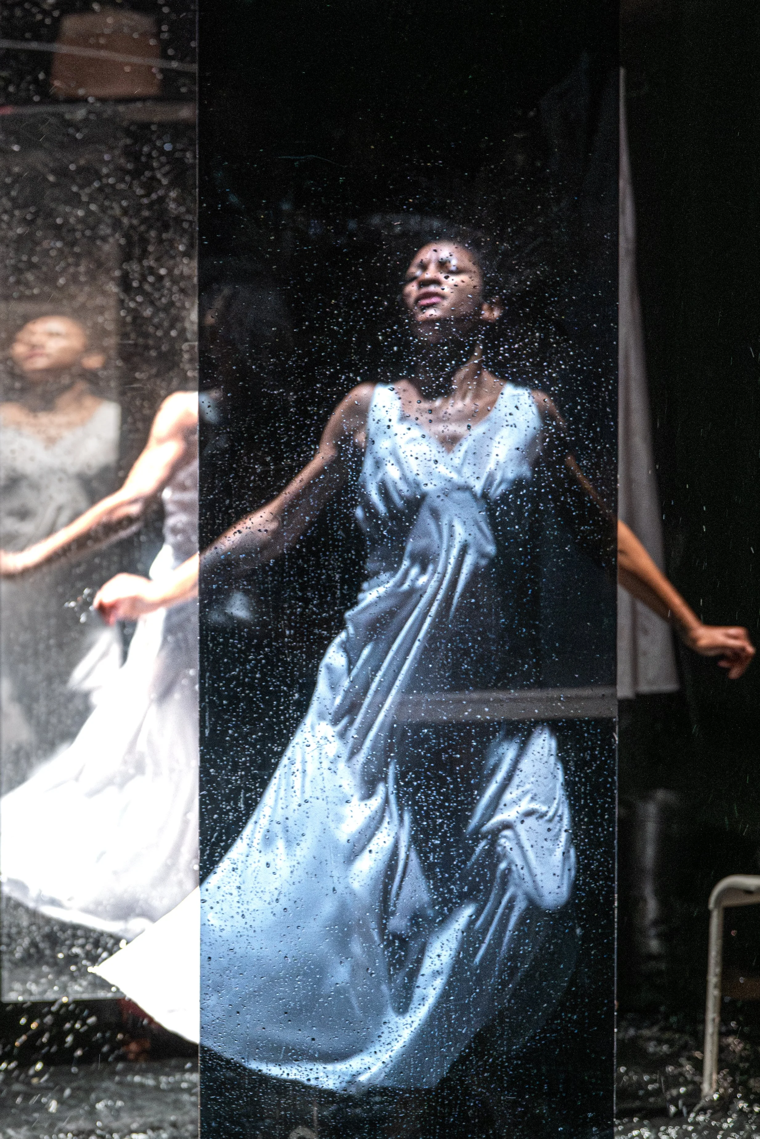 A woman in a silver dress is touching a wet, reflective surface, creating a distorted reflection of her face and upper body, with water droplets on the surface.