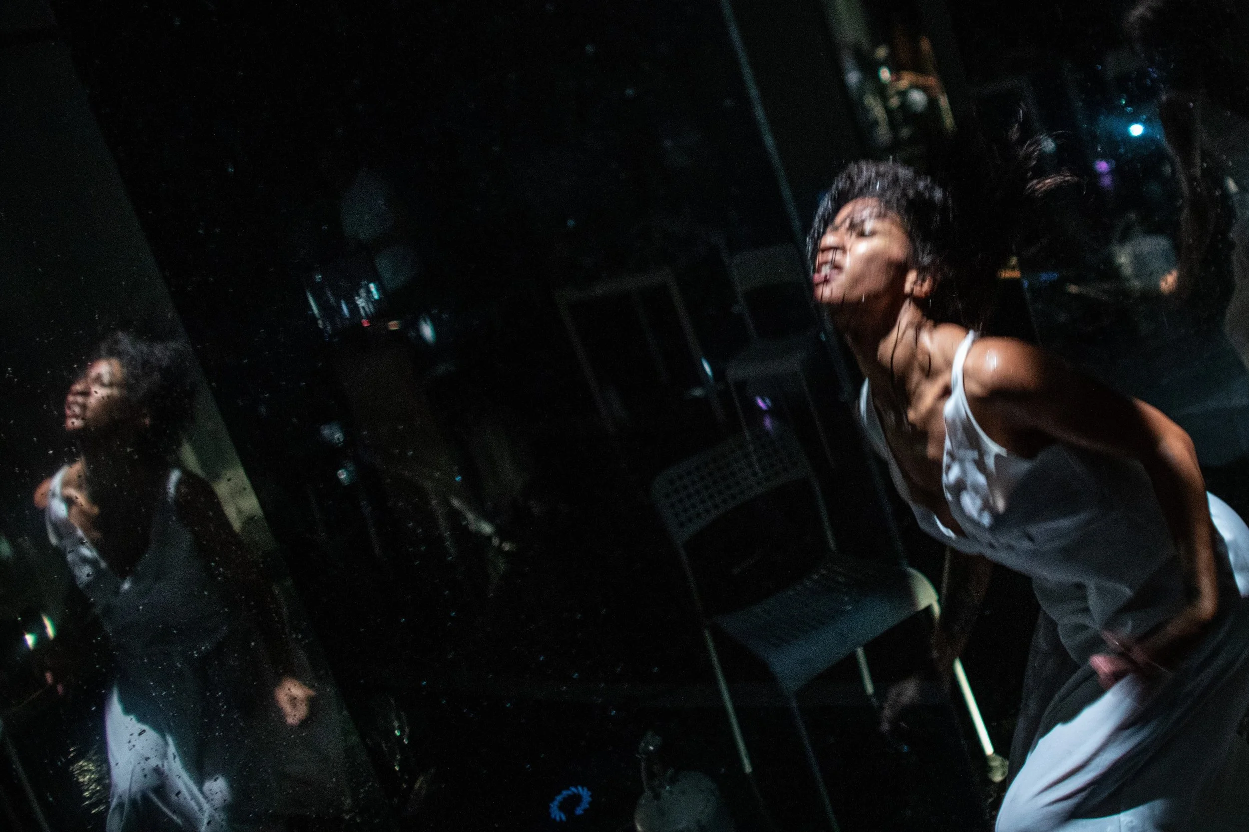 A woman with wet hair and a white sleeveless top is leaning against a mirror in a dark room, appearing to scream or yelp, with her eyes closed and her mouth open. Her reflection is visible in the mirror, and the room has black chairs and a metallic o