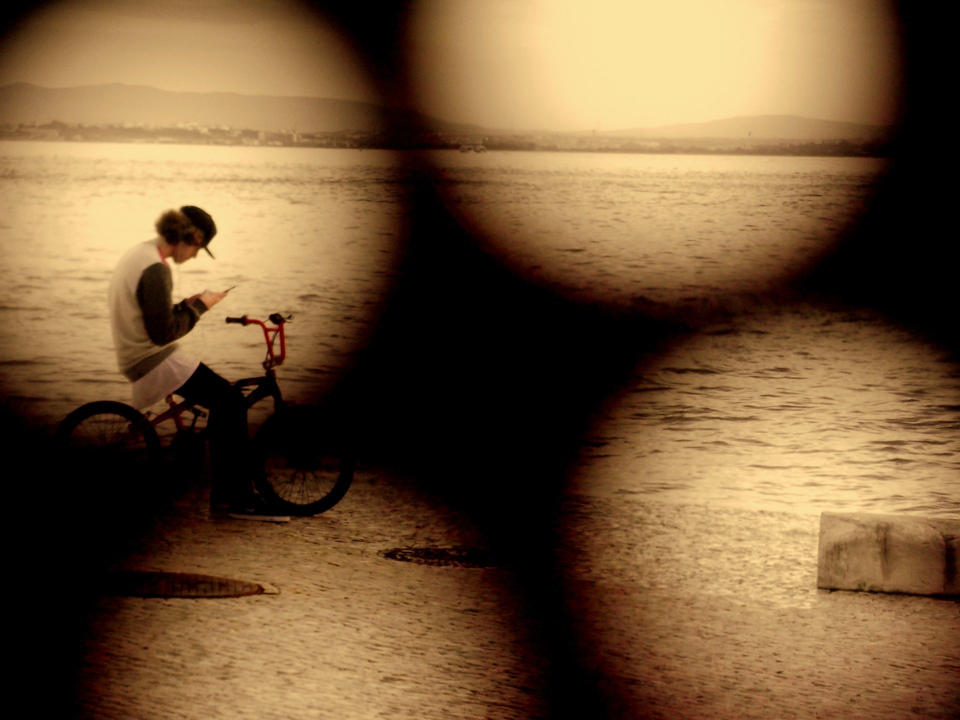 Person with curly hair and glasses standing next to a red bicycle, looking at their phone, near a body of water during sunset.