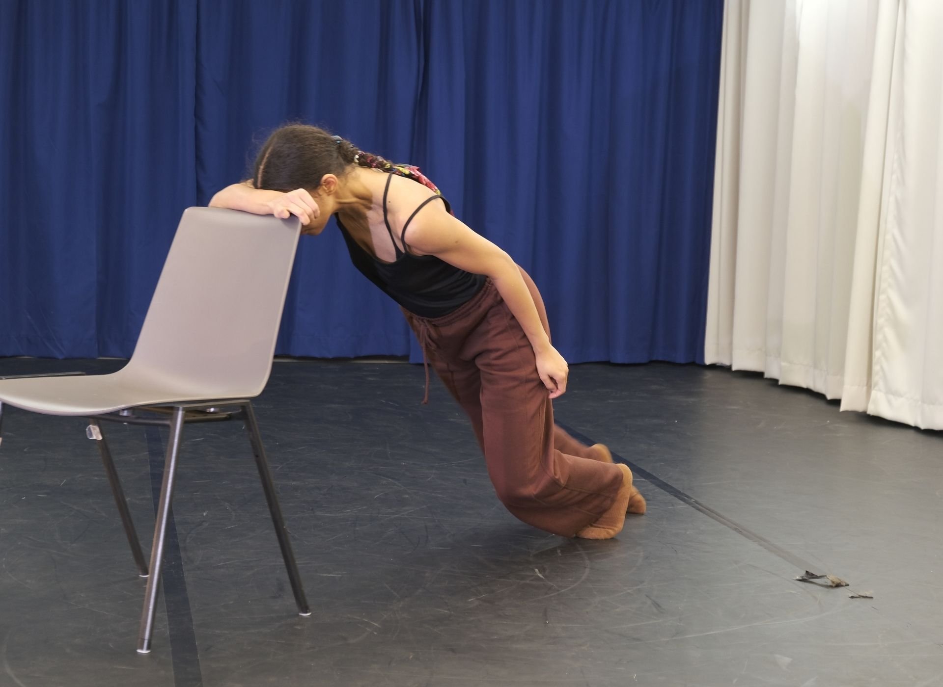 A woman is kneeling on the floor with her head against a chair, appearing to be exhausted or in despair, on a stage with blue curtains in the background.