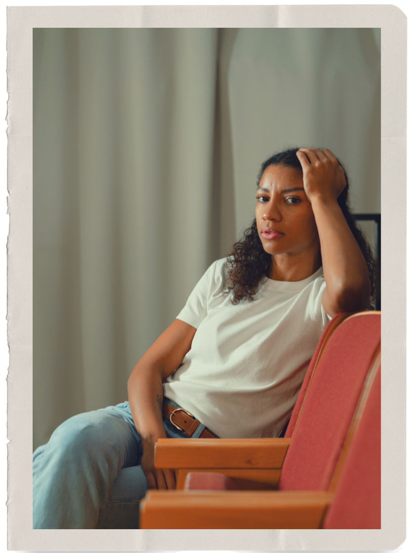 A woman sitting in a chair with a serious expression, one hand on her head, in front of beige curtains.