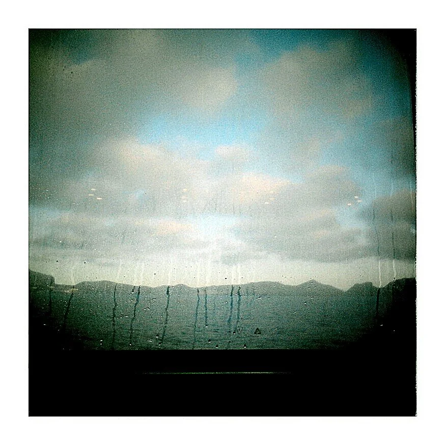 View of clouds over mountains seen through a window with rain droplets on it.