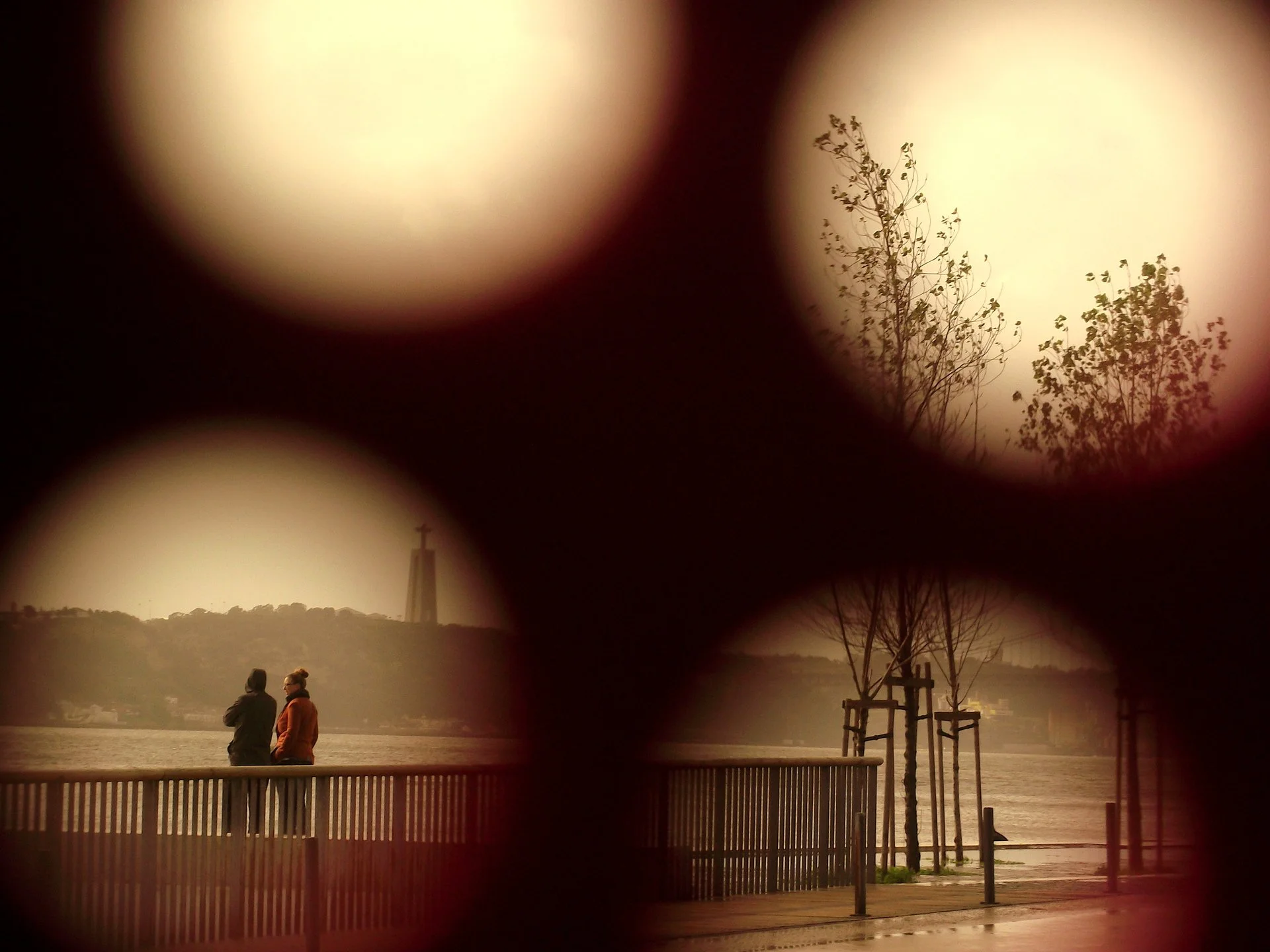 A photograph of two people standing on a waterfront promenade, viewed through a monocular telescope. Trees and a distant building are visible across the water, and the scene is taken during the evening or early morning with a warm, subdued light.
