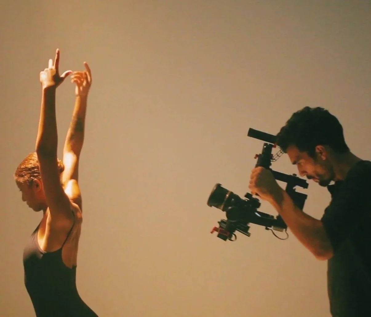 A woman with short hair wearing a black tank top is stretching her arms above her head while a man with dark hair operating a camera with a stabilizer captures her in action.