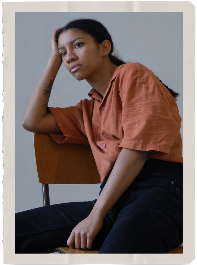 A young woman with dark hair, wearing a rust-colored shirt and black pants, sitting on a wooden chair with a gray background, resting her head on her left hand with a contemplative expression.