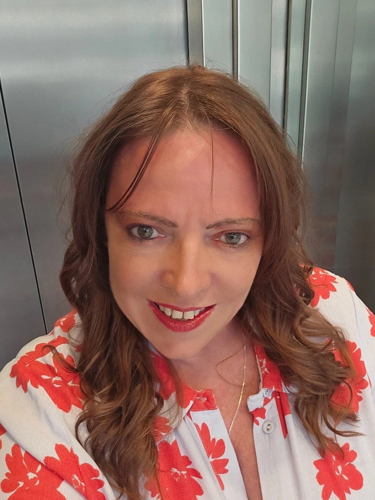 A woman with long, wavy brown hair and light-colored eyes taking a selfie in an elevator. She is wearing a white blouse with red floral patterns and smiling.