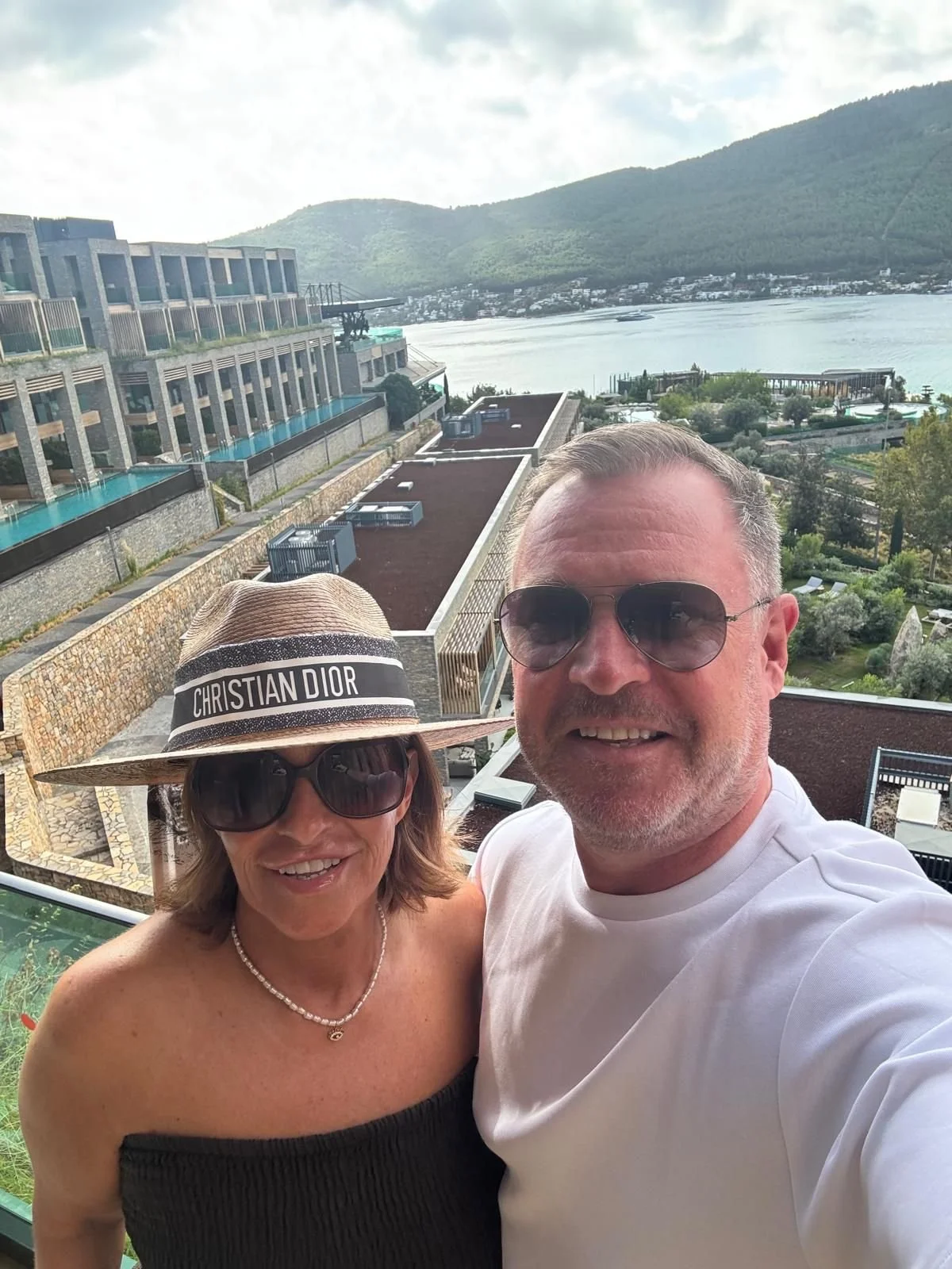 A smiling man and woman taking a selfie on a balcony overlooking a lake and mountains. The woman is wearing a Christian Dior sun hat and sunglasses. The man is wearing sunglasses and a white t-shirt.